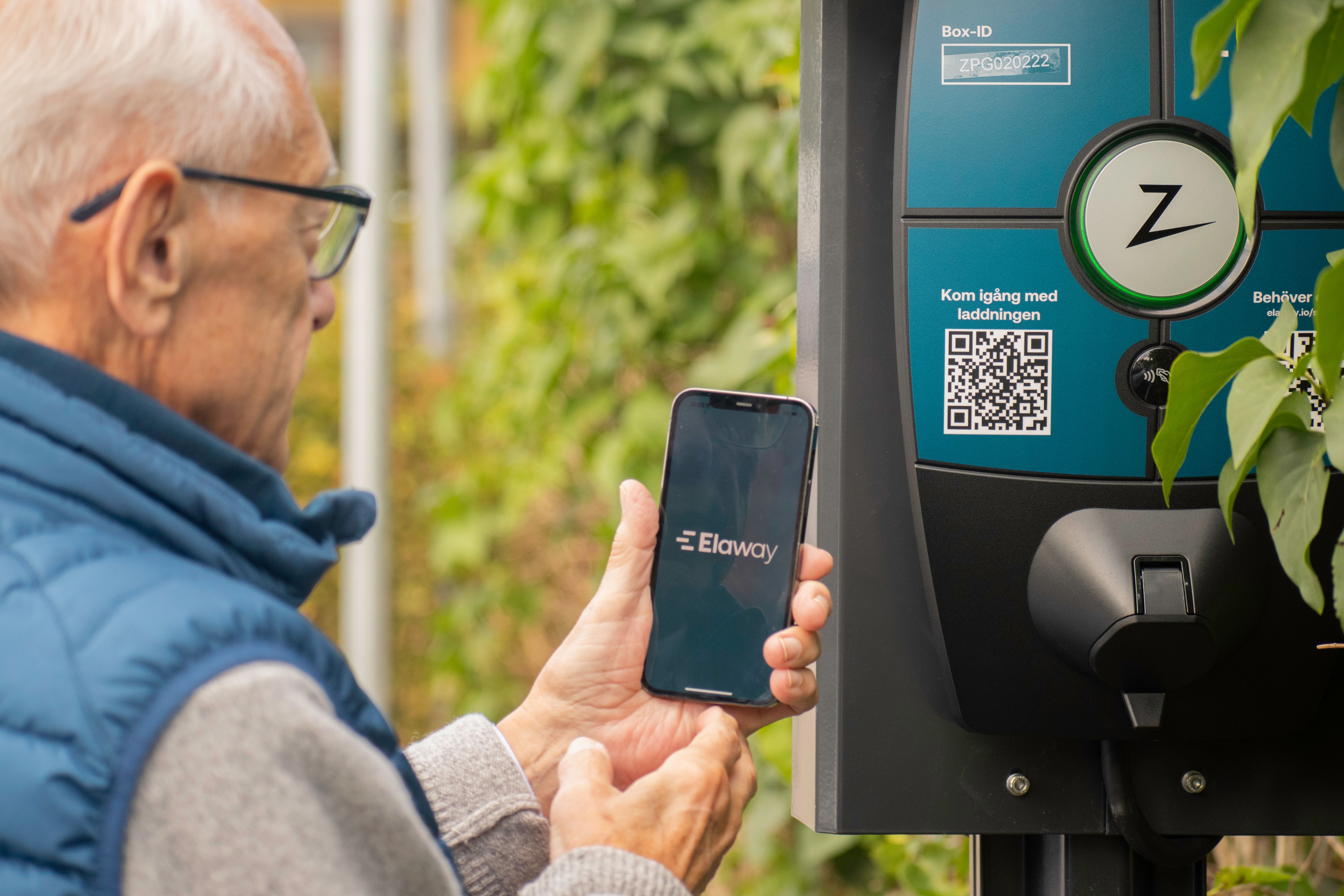 bilde av en eldre mann som holder en telefon foran en ladeboks for elbil.
