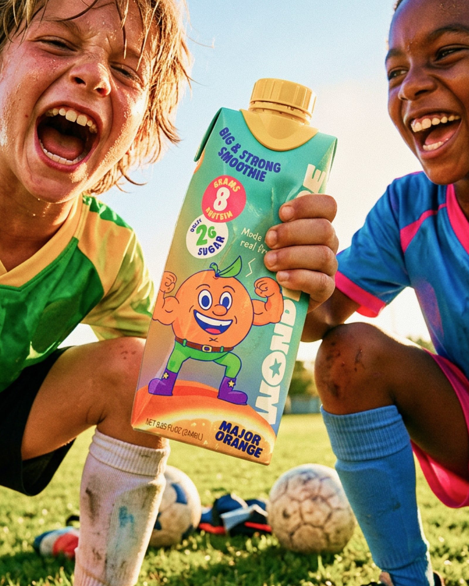 Two laughing kids in soccer kits crouch on a field, one holding a Major Orange smoothie toward the camera.