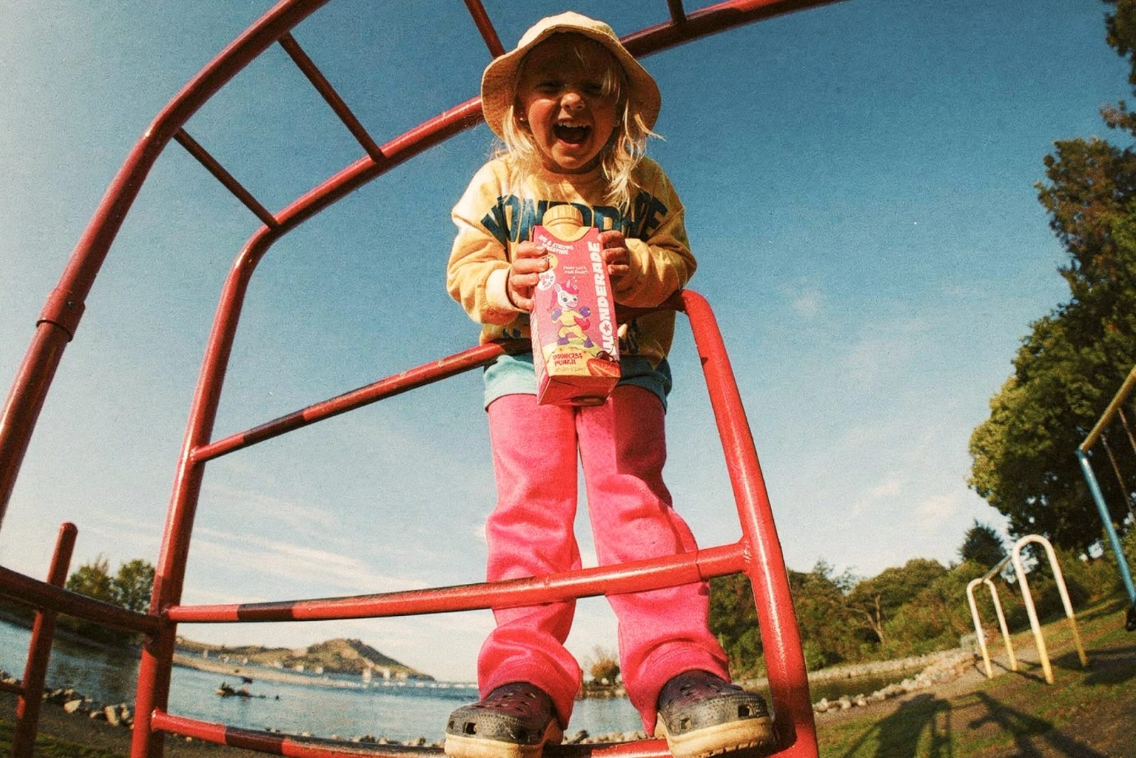 A laughing child in a Wonderade sweatshirt stands on a red climbing frame by the water, holding a Princess Punch carton.