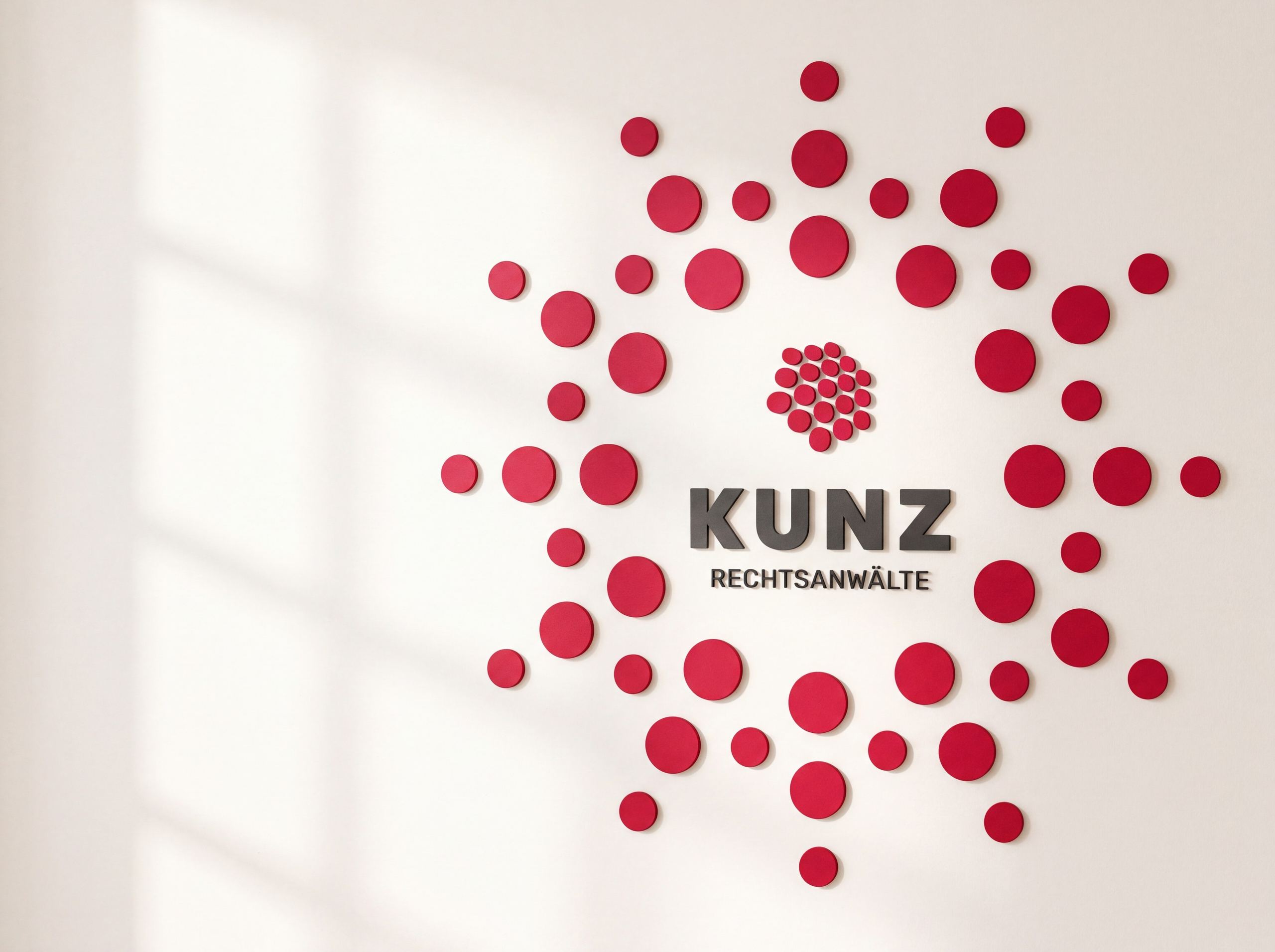 Rote Beeren des KUNZ-Logos als Ring an einer hellen Wand, in der Mitte das Logo