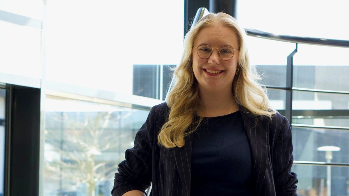 Smilende person med briller står foran store vinduer i et lyst lokale