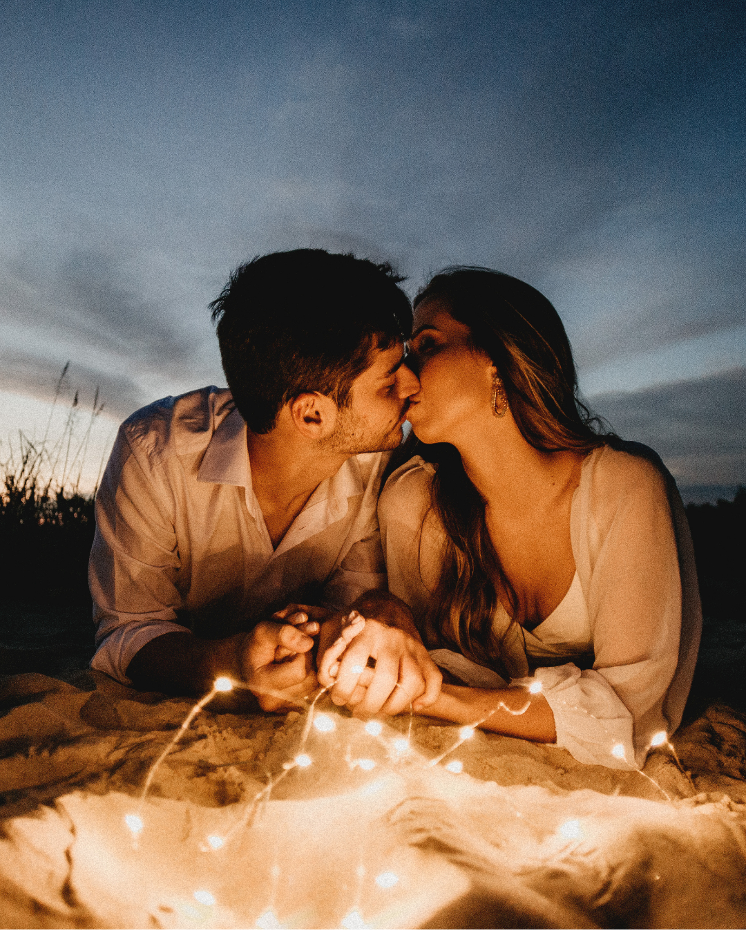 A couple exchanging a kiss on a beach at dusk, surrounded by twinkling lights.