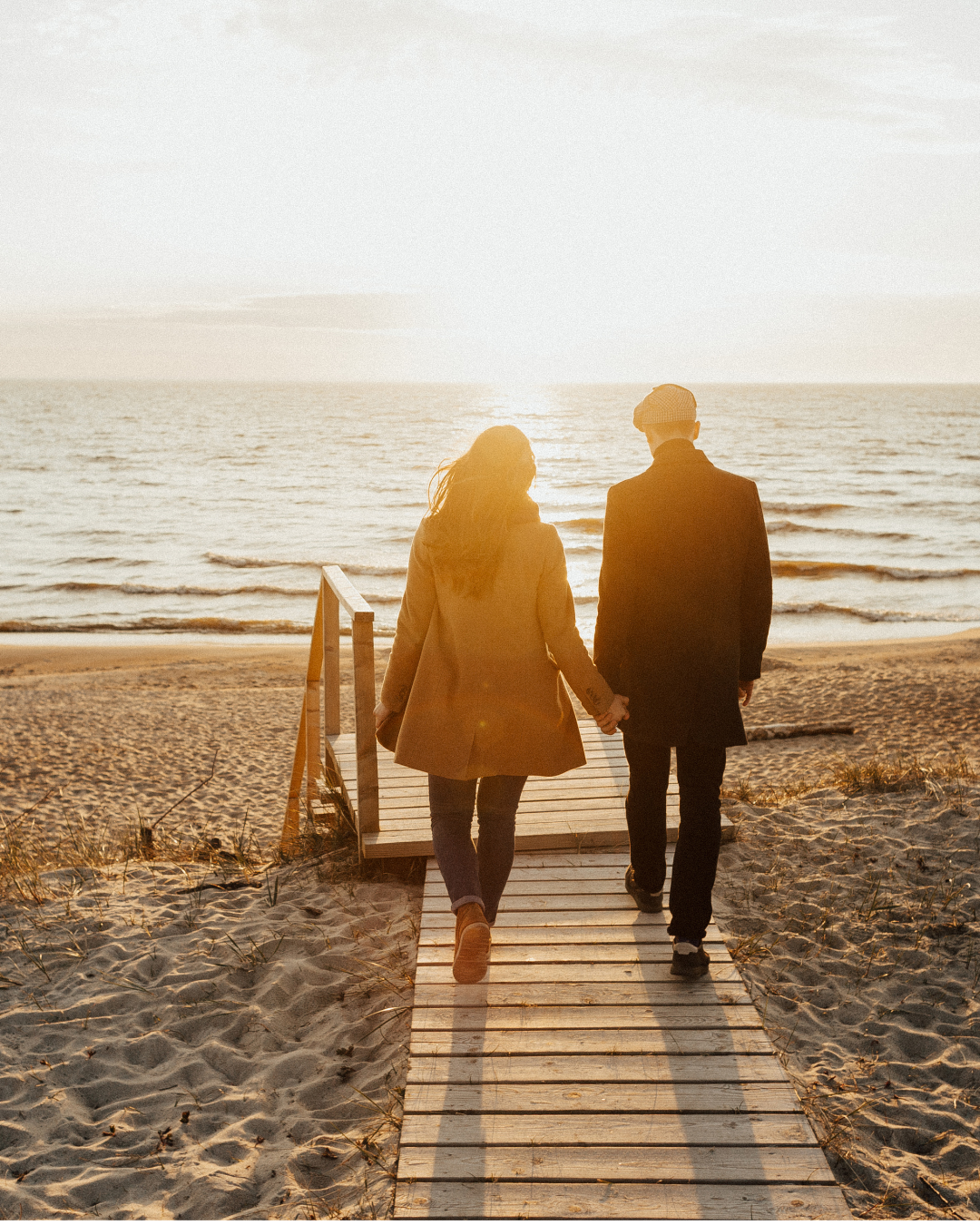 couple holding hands and walking on a beach boardwalk towards the ocean at sunset