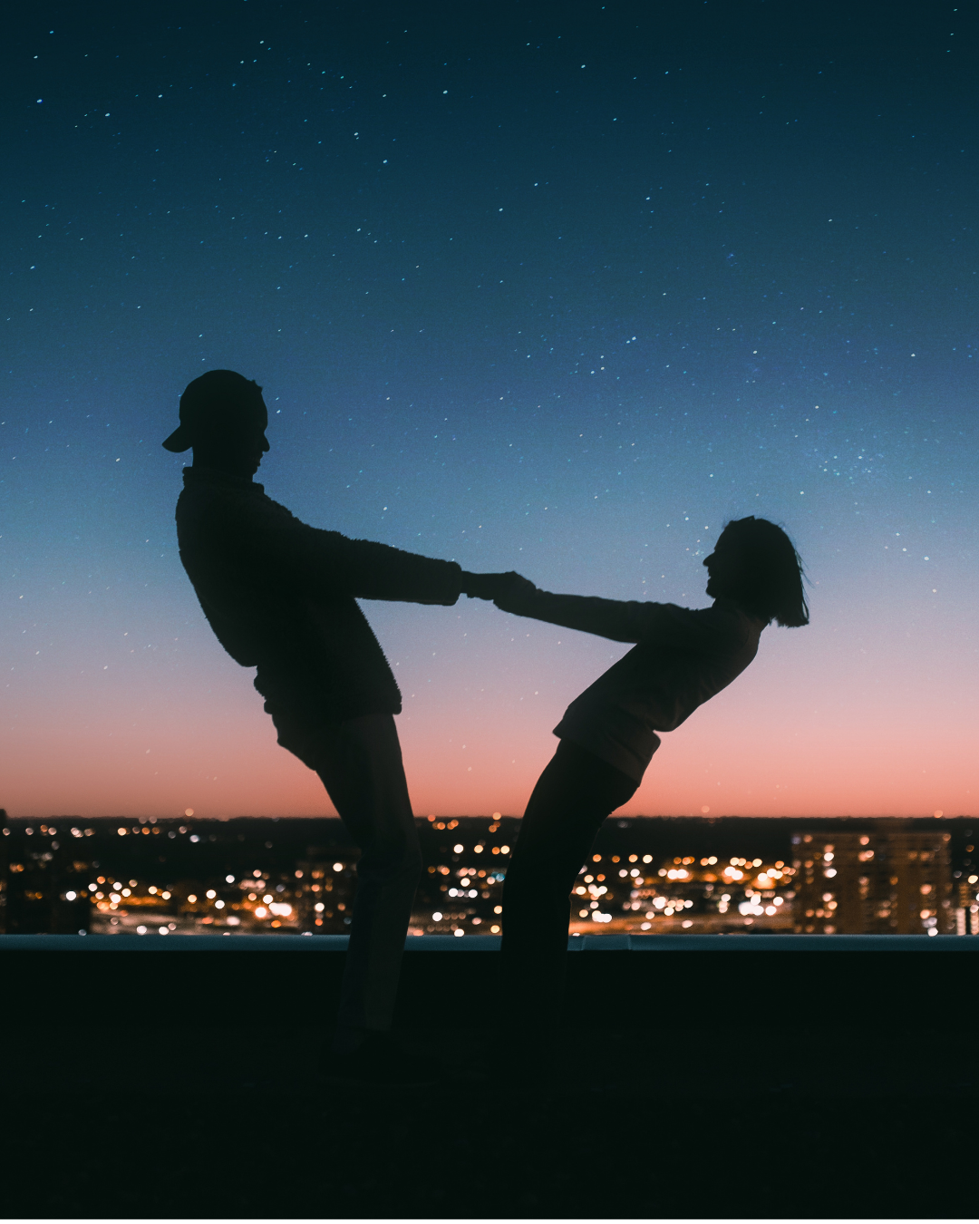 couple holds hands while leaning backward as they are shadowed by a sunset backdrop of the city at night