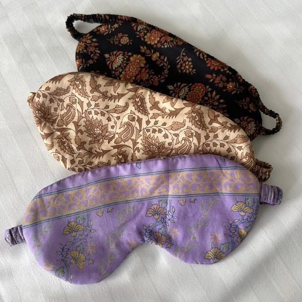 Four fabric sleep masks stacked, featuring floral and patterned designs in brown, cream, and purple.