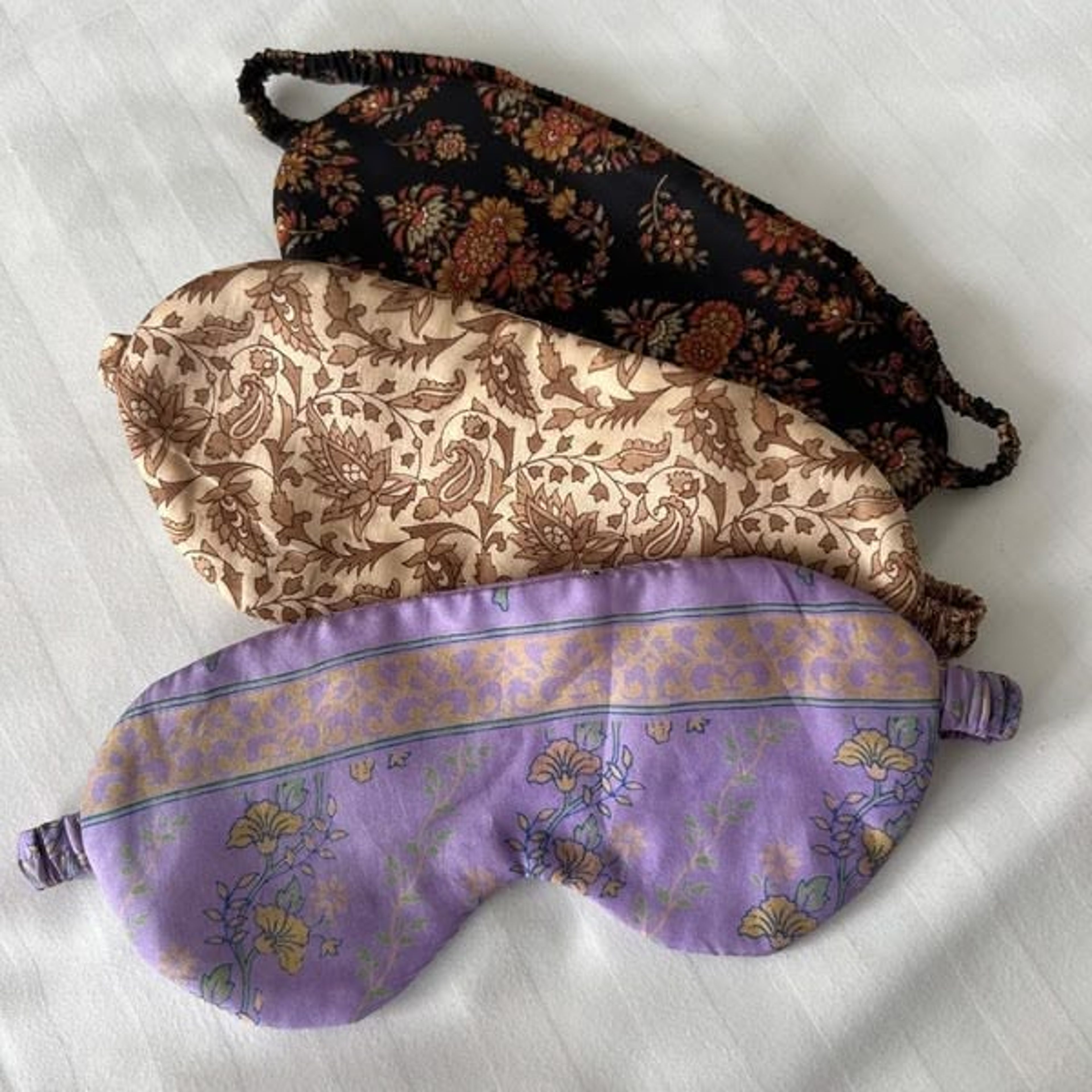 Four fabric sleep masks stacked, featuring floral and patterned designs in brown, cream, and purple.