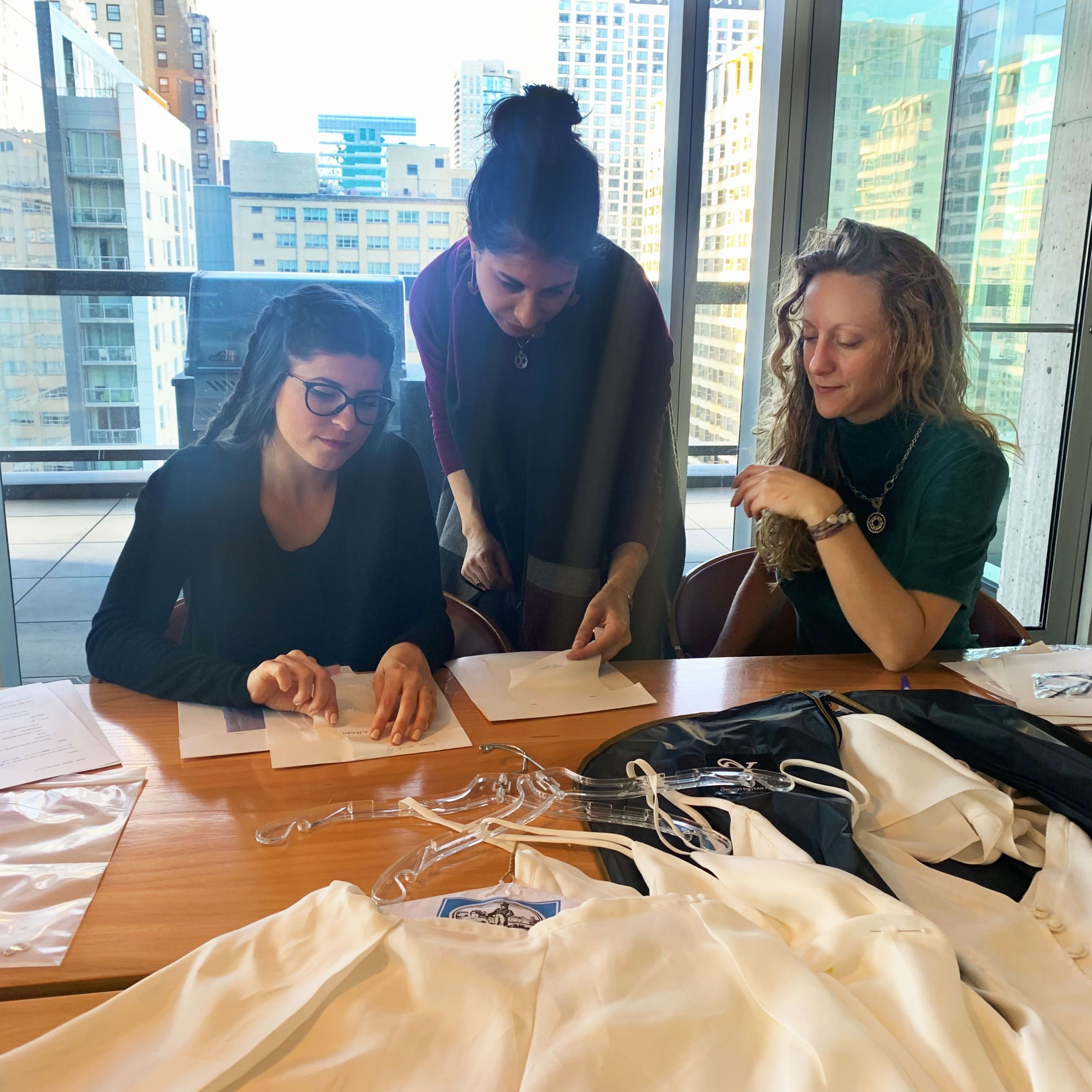 Three women examine clothing items at a high-rise office table with city skyline visible behind them.