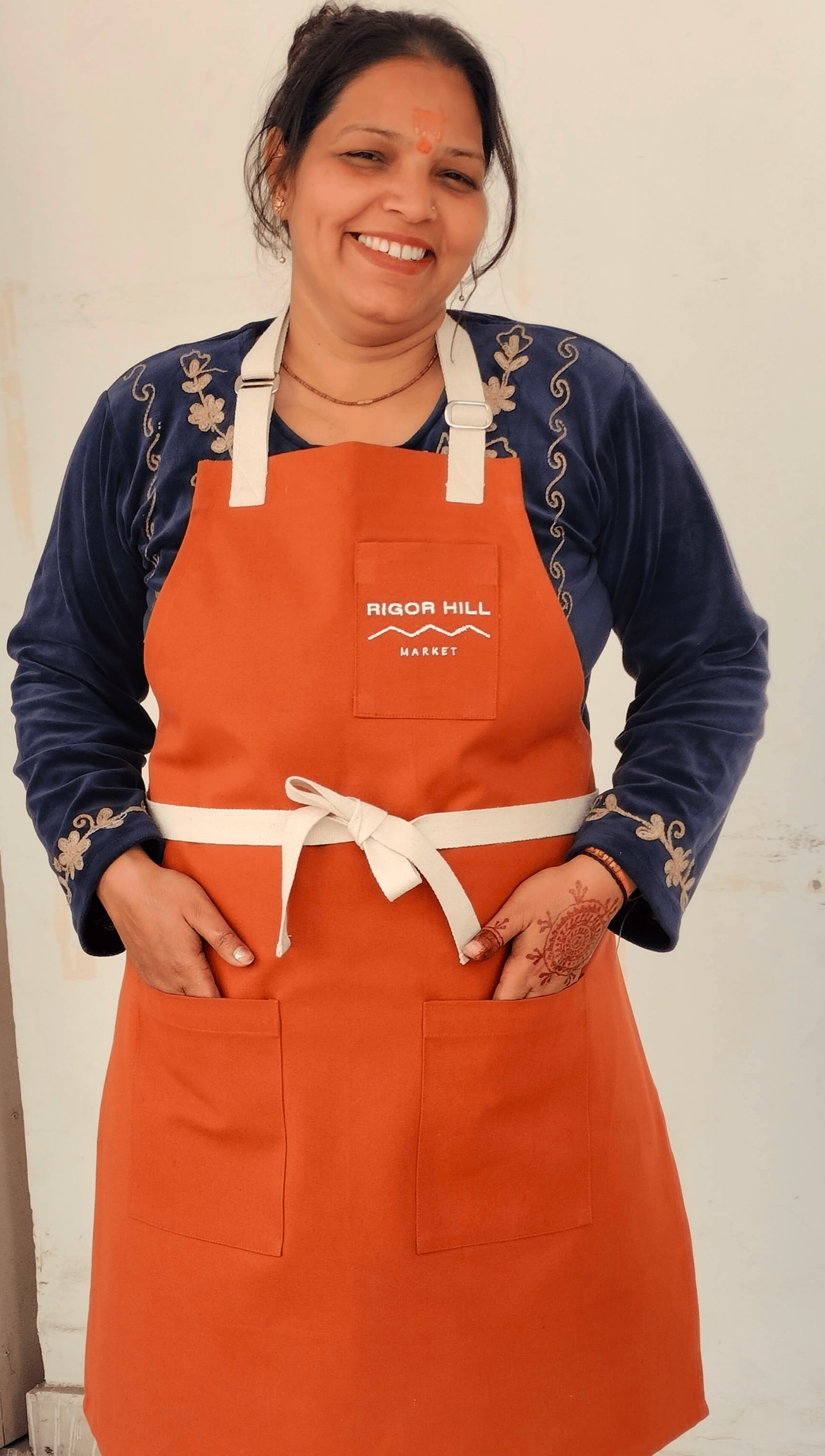 Woman smiling wearing orange Rigor Hill Market apron over navy embroidered jacket.