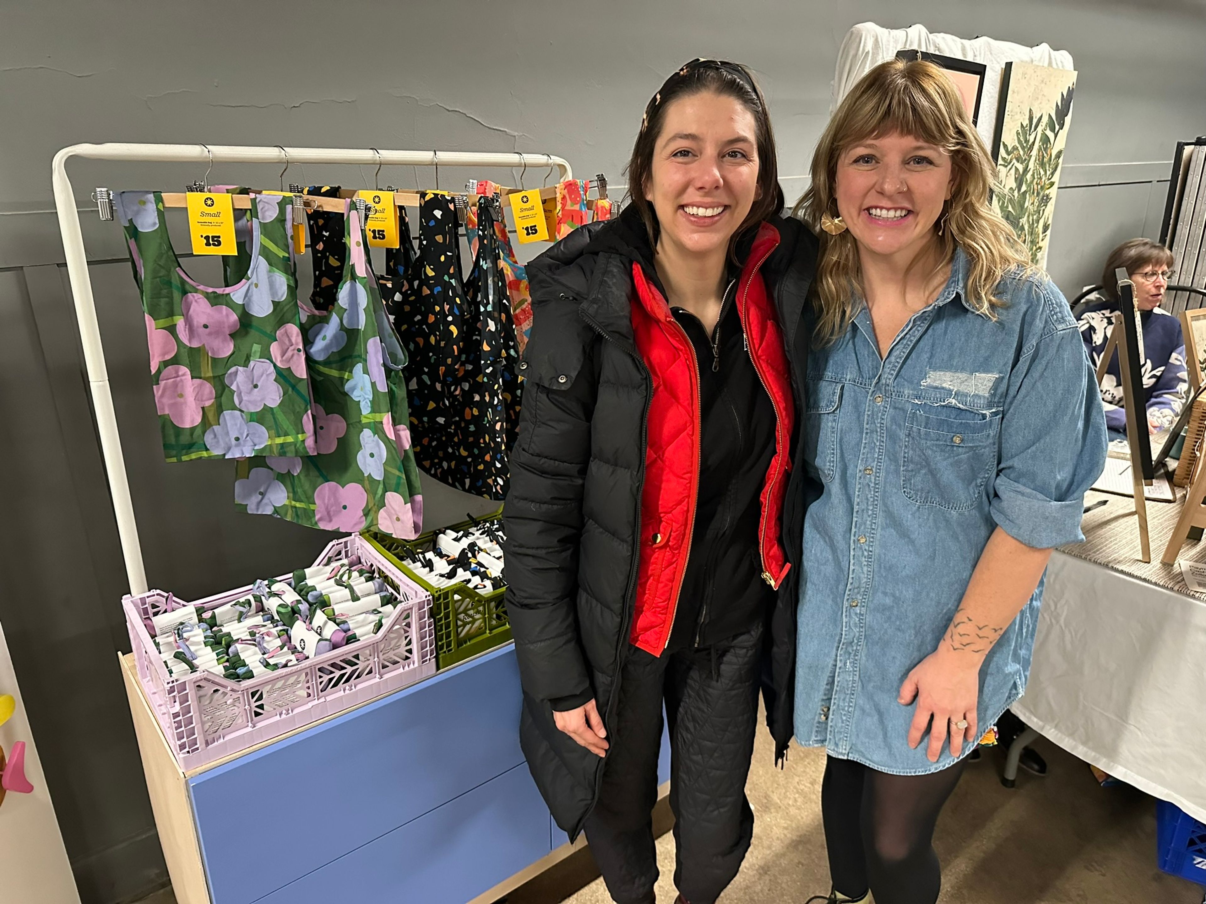 Two women smiling at retail clothing display with floral and patterned garments priced at $15.