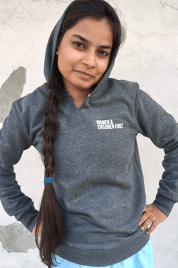 Young person wearing gray hoodie with long dark hair, hand on hip, indoors.