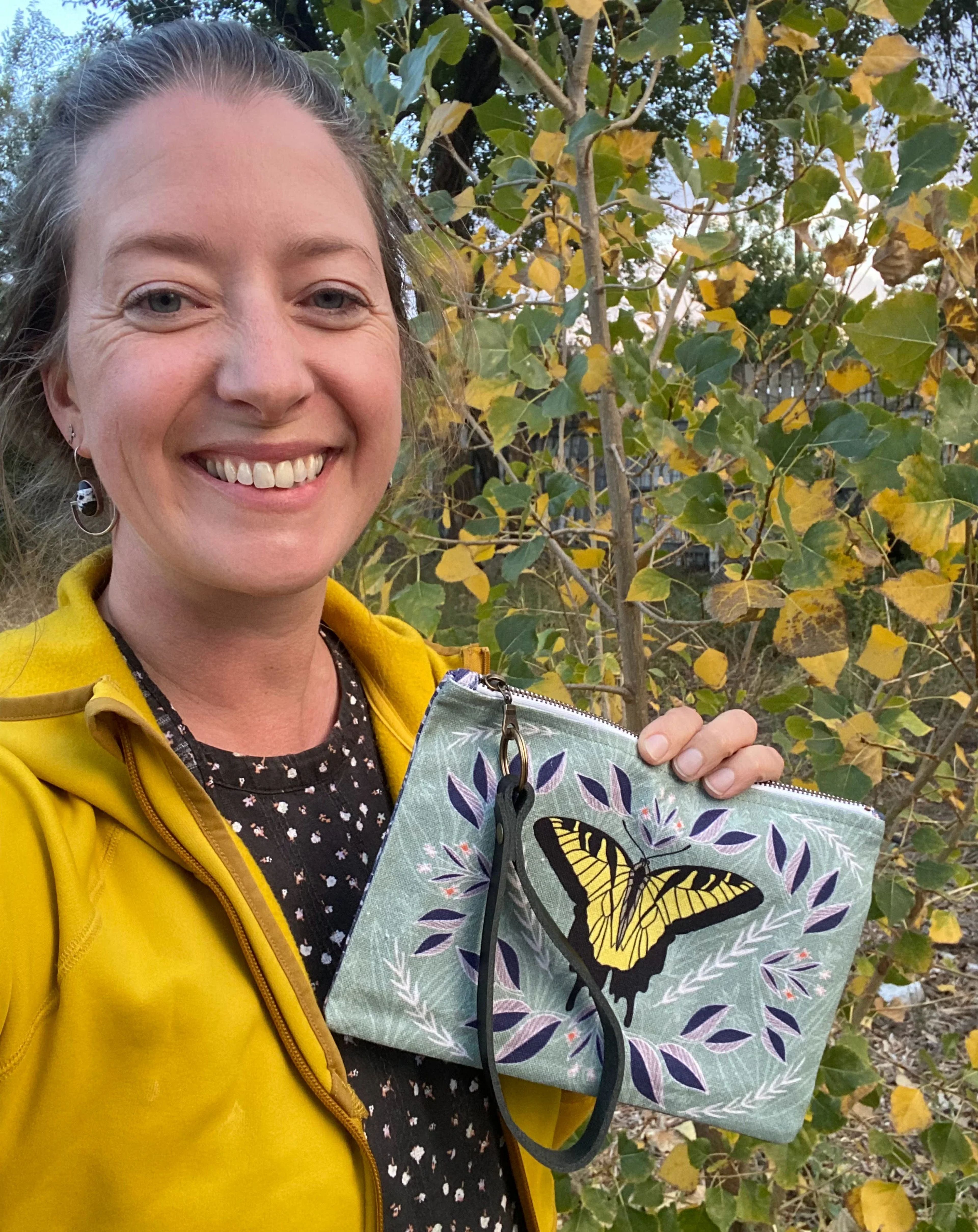 Woman in yellow jacket holding butterfly-patterned pouch outdoors among autumn foliage.