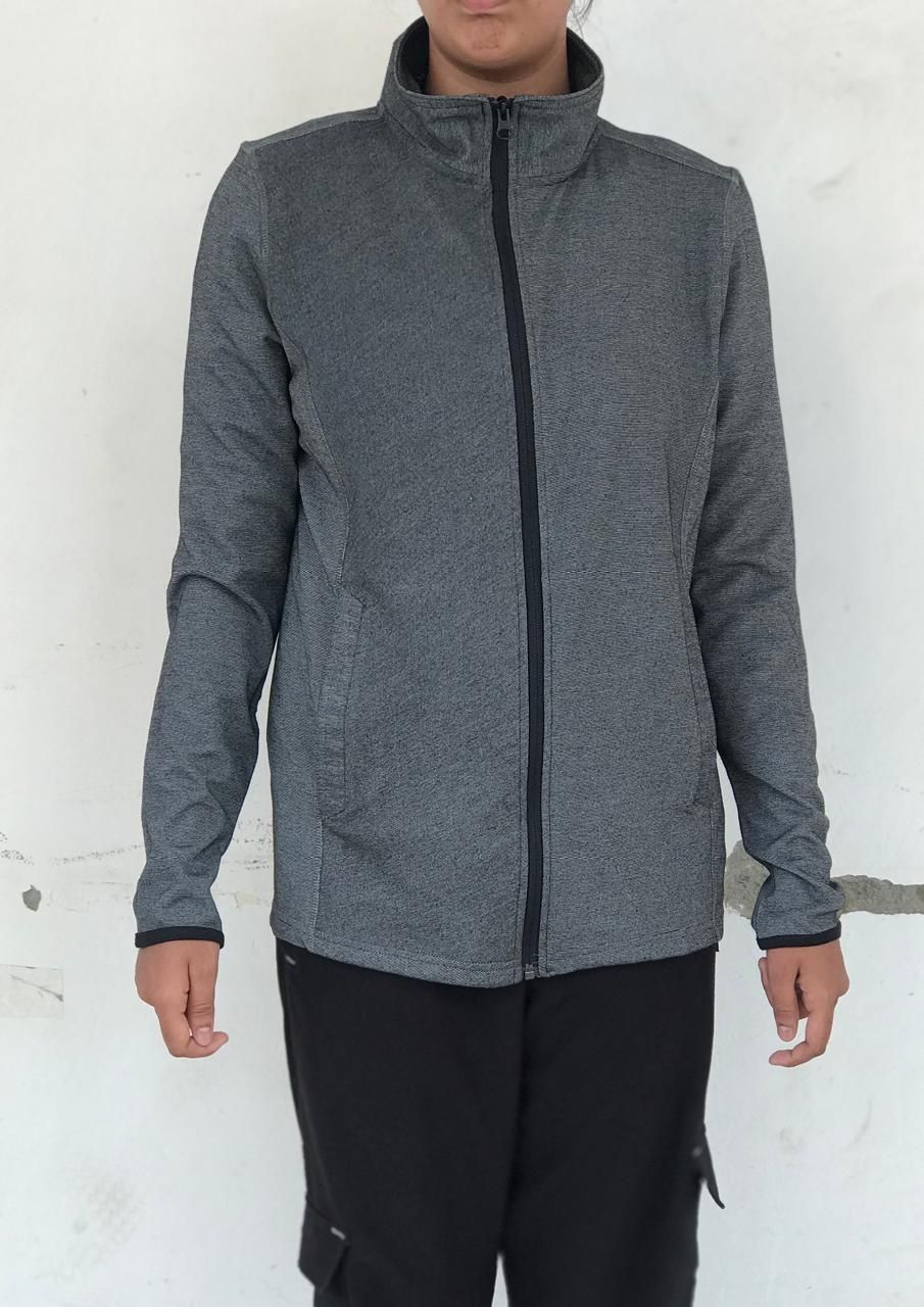 Person wearing gray zip-up fleece jacket and black pants against white wall.