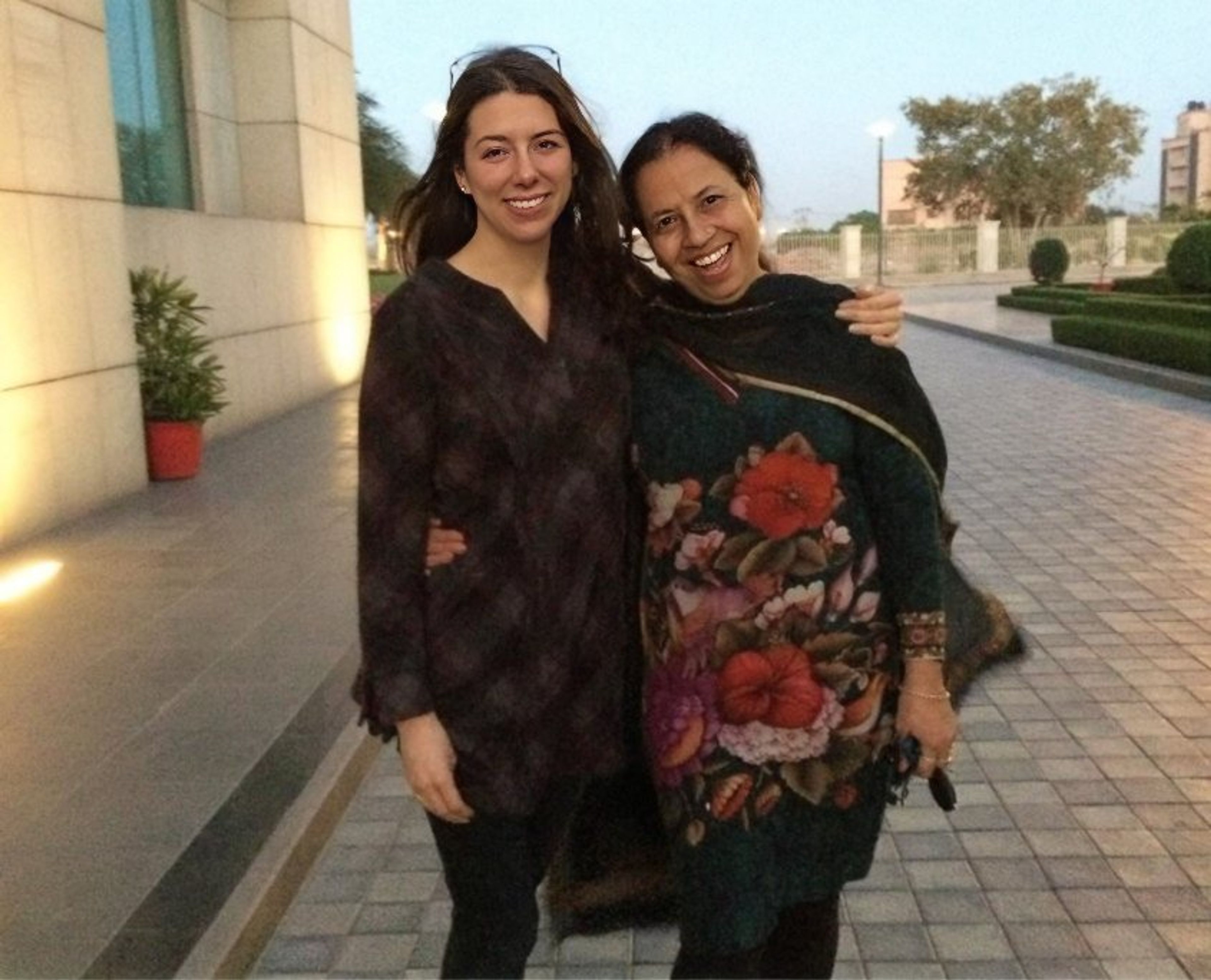 Two smiling women posing together on a paved outdoor pathway near modern buildings.