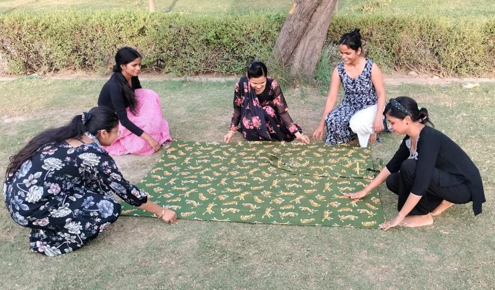 Five women sit in circle on grass around green patterned fabric outdoors.