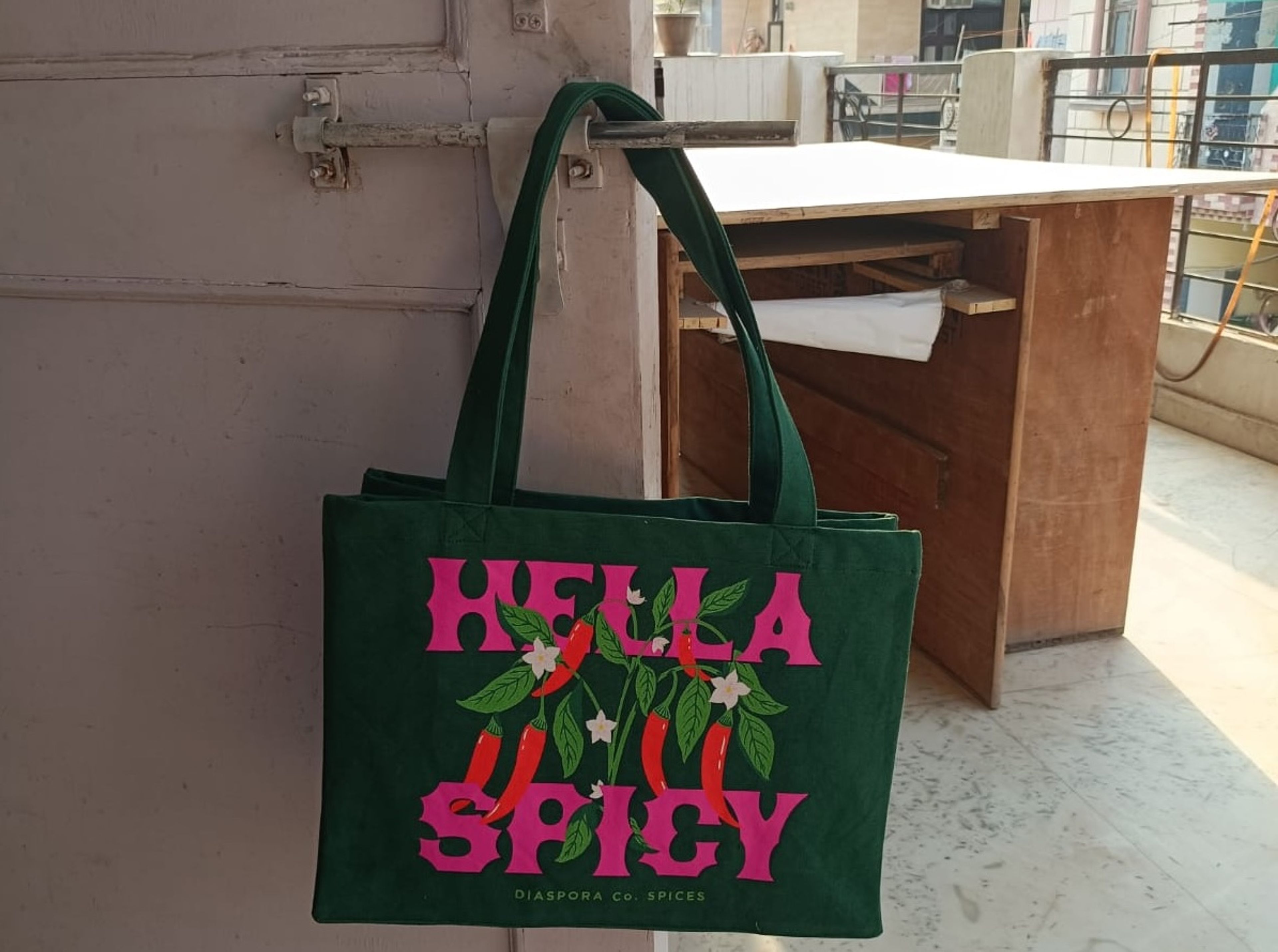 Green tote bag with "Hella Spicy" text and chili pepper design in warehouse setting.