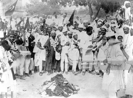 # Alt text:

Large crowd of men in traditional dress gathered around fire, holding flags, historical photograph.