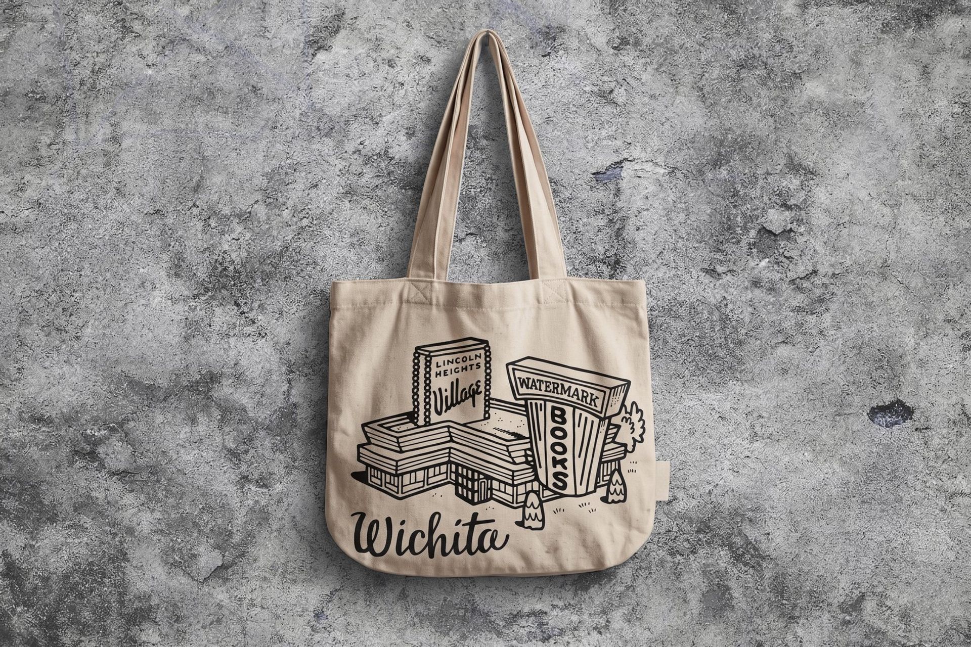 A canvas tote bag hanging on a weathered concrete wall, featuring black line-art illustrations of the Watermark Books & Cafe and Lincoln Heights Village buildings above the word "Wichita" in script.