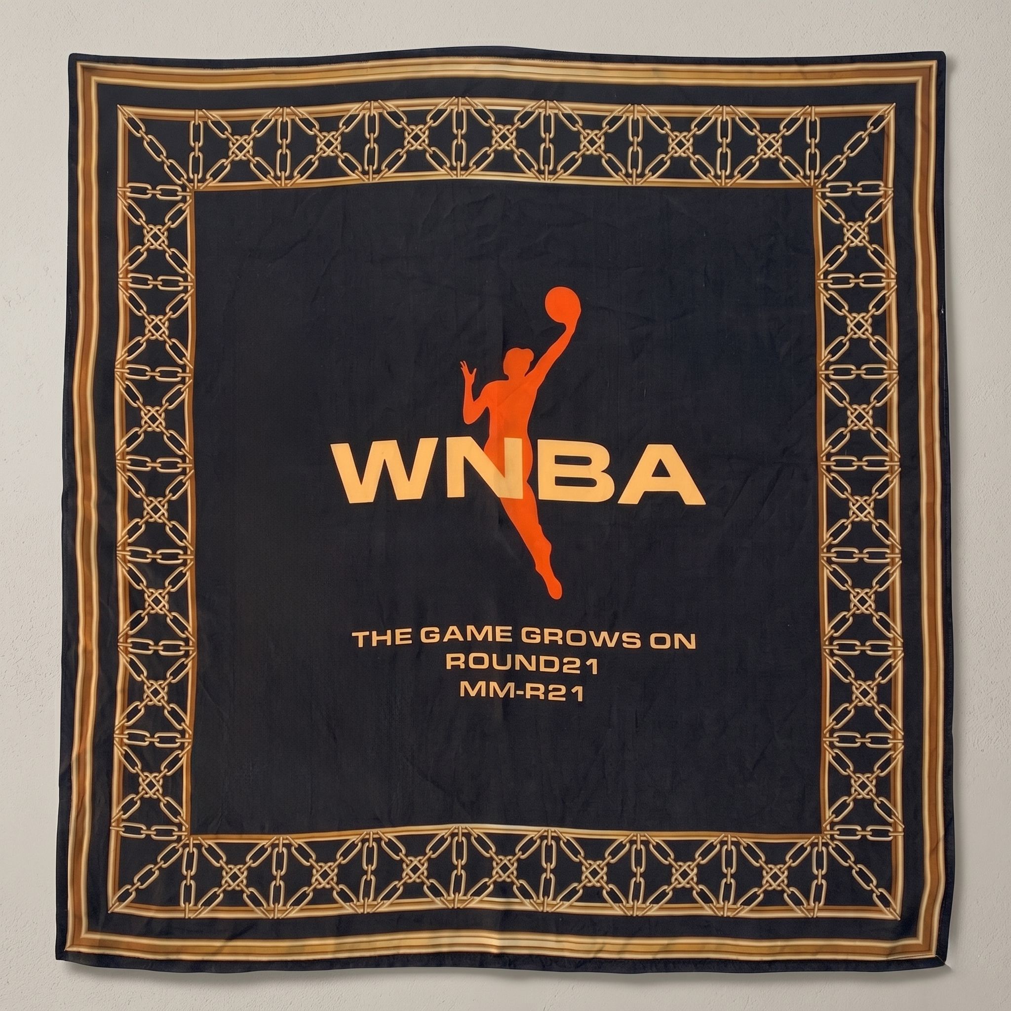 "Black silk bandana featuring an orange WNBA logo and player silhouette in the center. The design is framed by an intricate gold chain-link border and includes the text 'The Game Grows On' at the bottom."