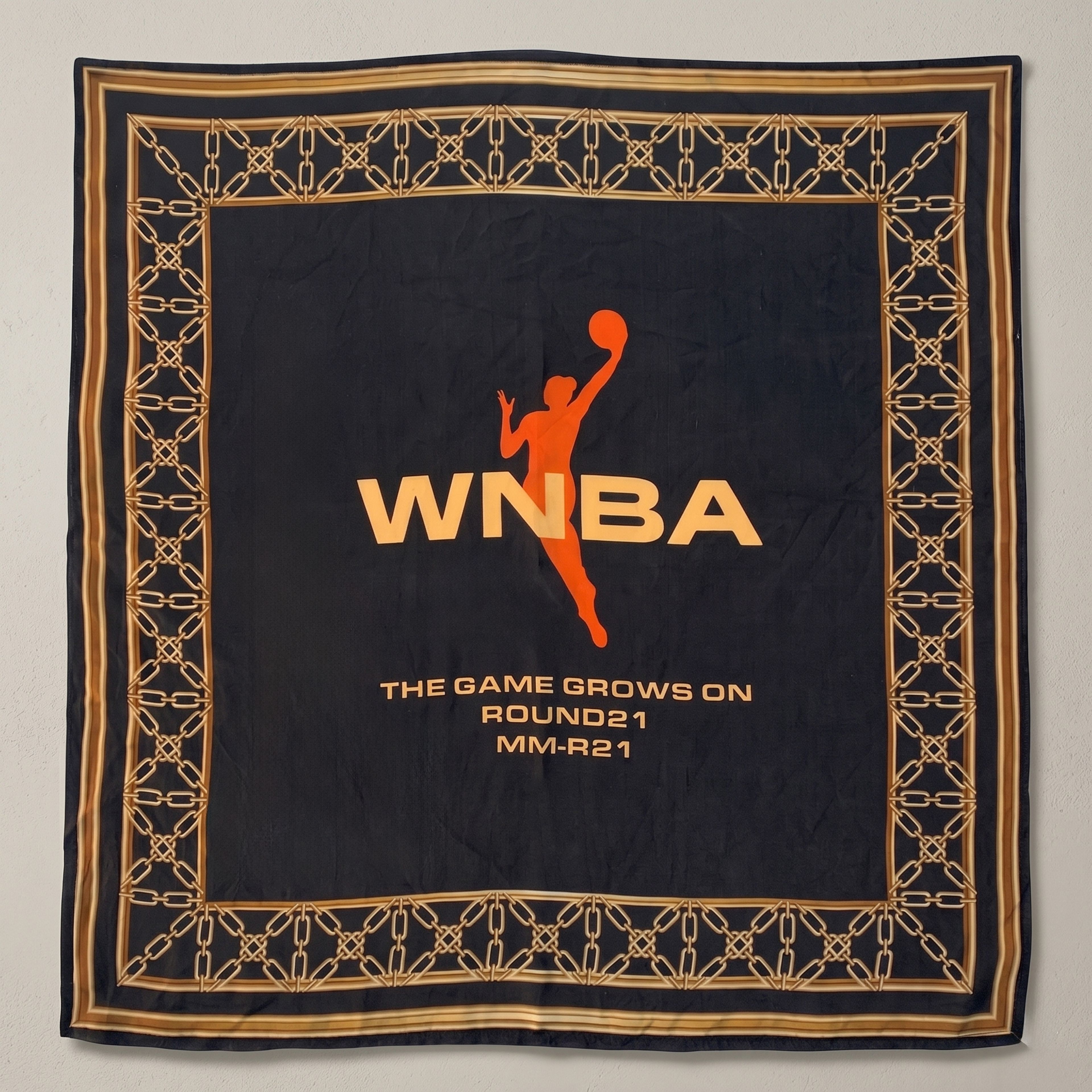 "Black silk bandana featuring an orange WNBA logo and player silhouette in the center. The design is framed by an intricate gold chain-link border and includes the text 'The Game Grows On' at the bottom."