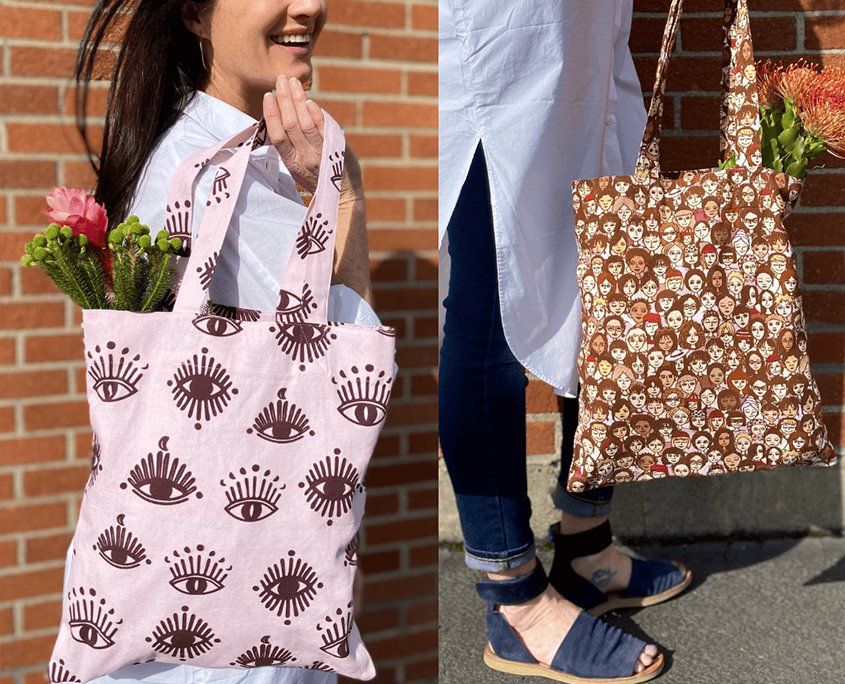 Two tote bags displayed outdoors against brick wall: eye-patterned pink and skull-patterned brown.