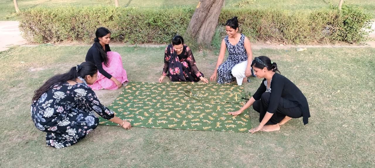 Five women sitting in circle on grass, spreading out patterned green fabric outdoors.