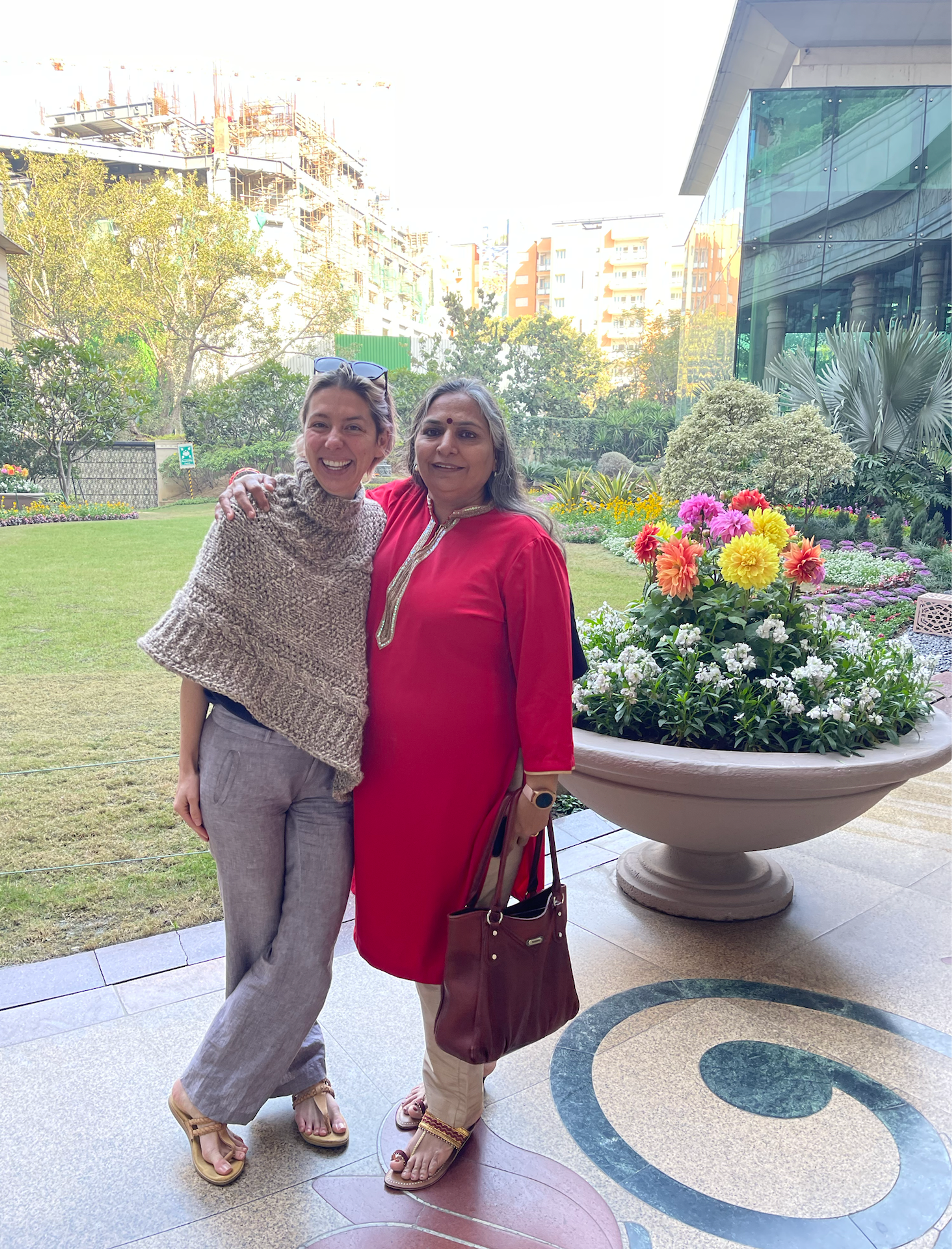 Two women smiling in a garden courtyard with colorful flowers and urban buildings behind them.