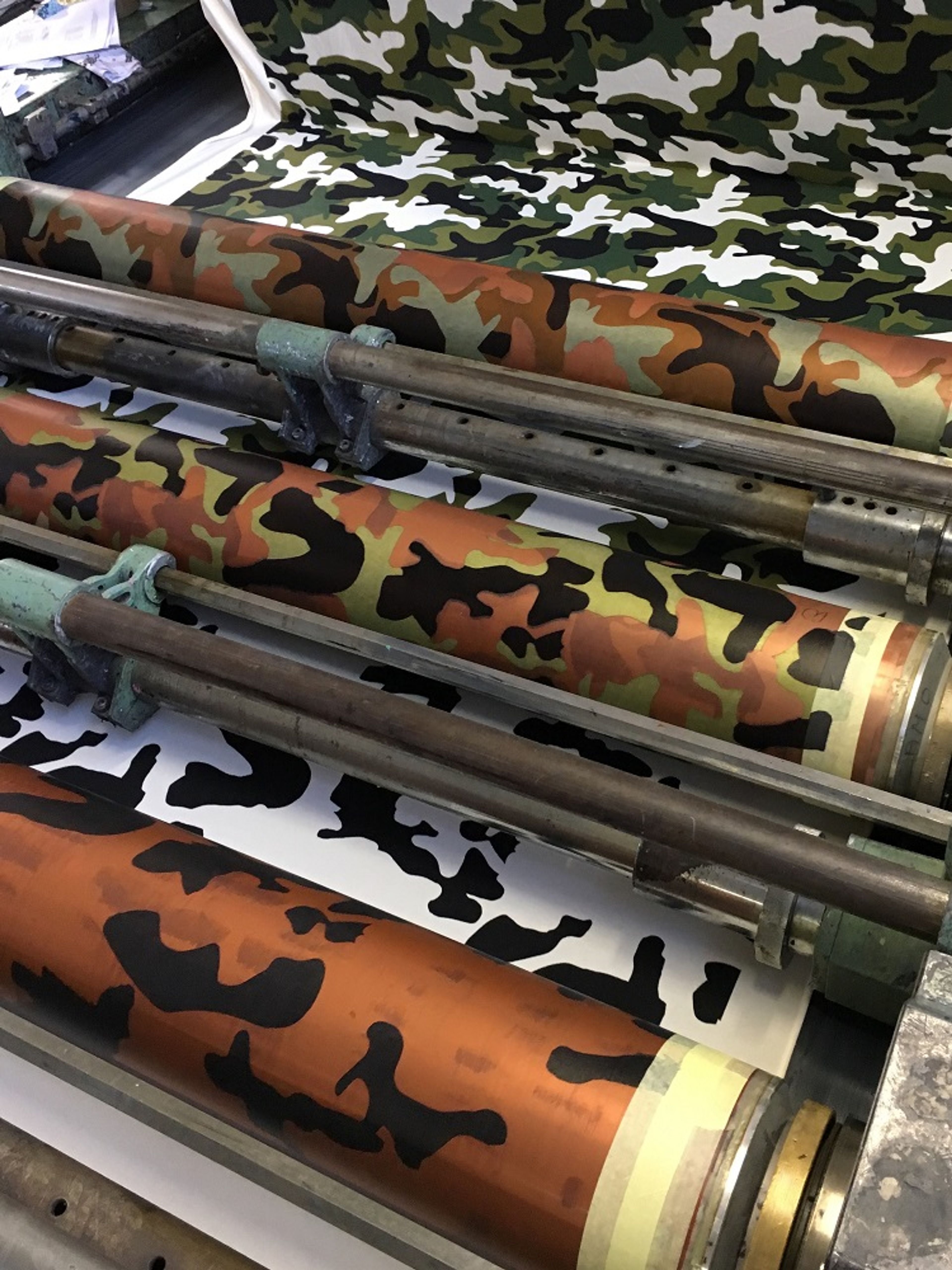 Rolls of camouflage patterned vinyl in various colors stacked on printing press rollers.