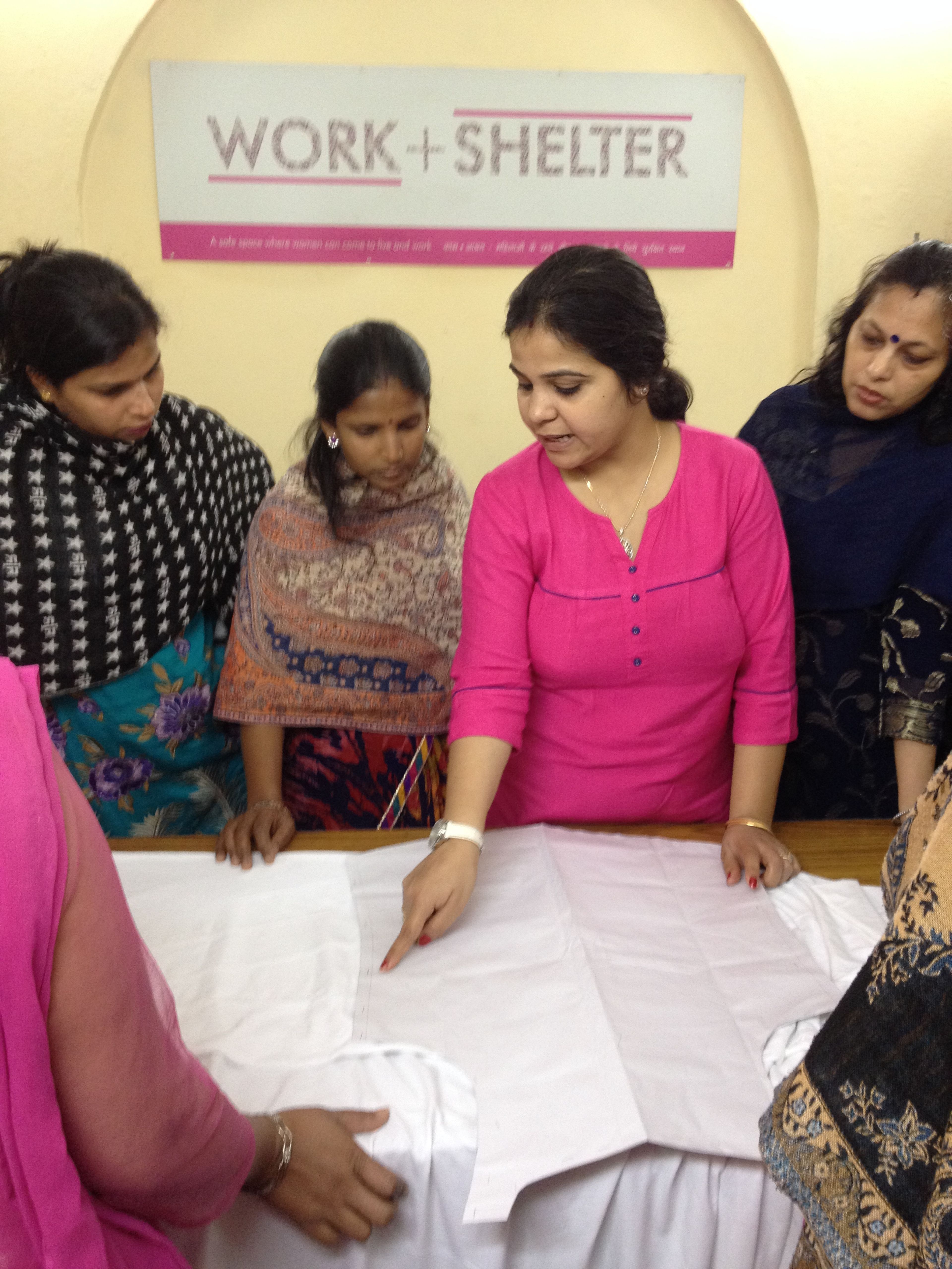 Women gathered around table at Work + Shelter organization, examining white fabric or documents together.