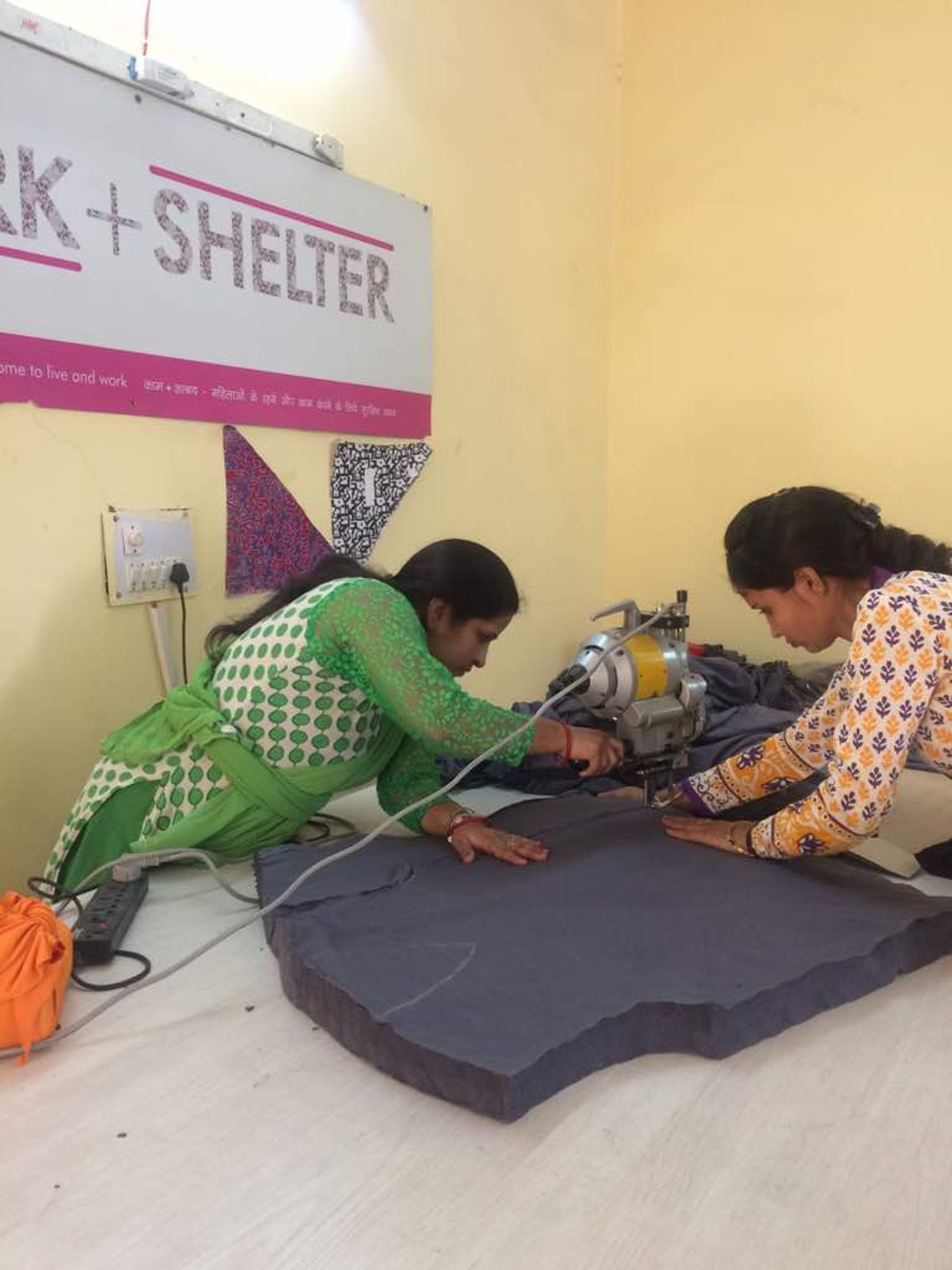 Two women operating sewing machine on dark fabric in vocational training shelter.