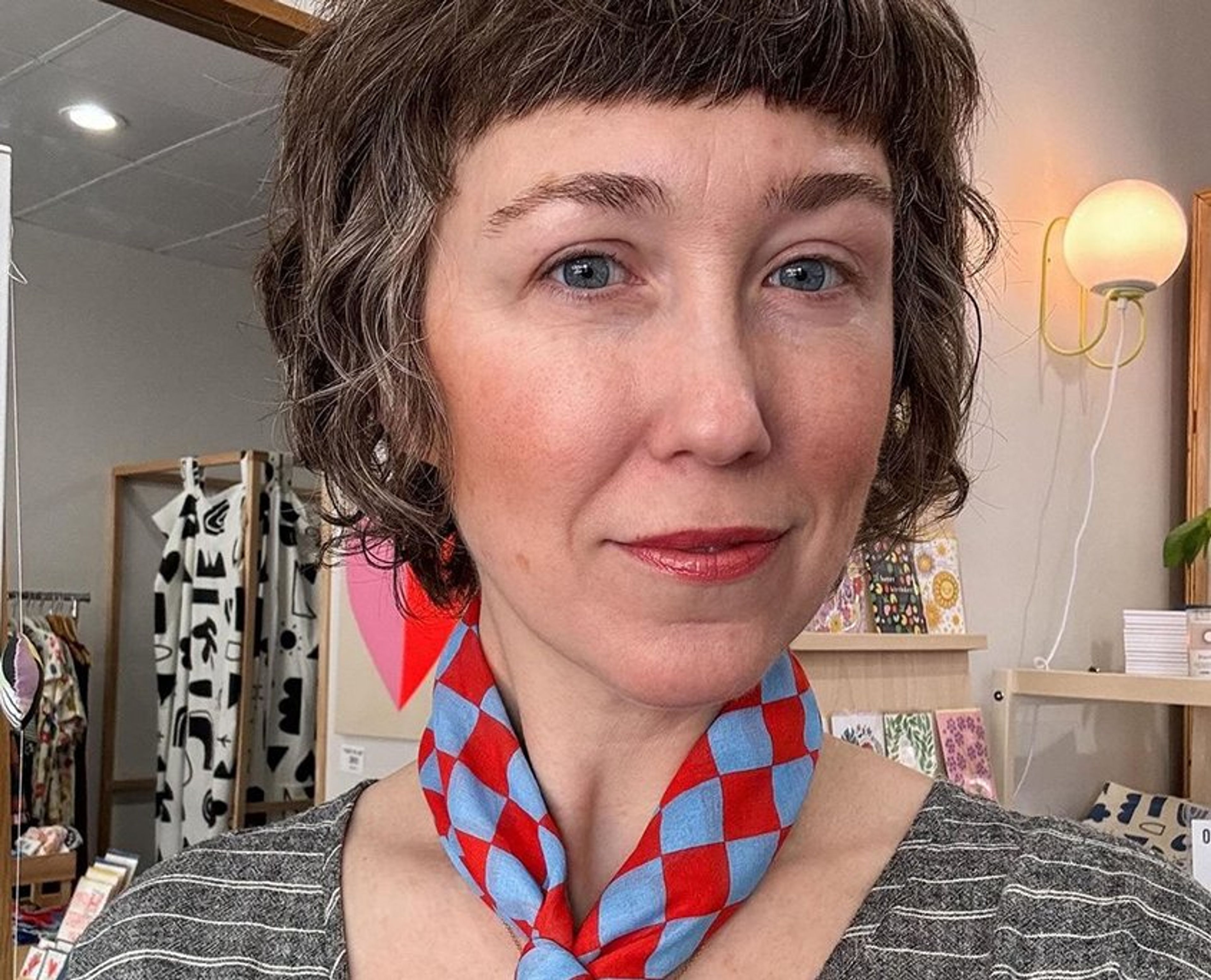 Woman with dark wavy hair and red lipstick wearing red and blue checkered scarf indoors.