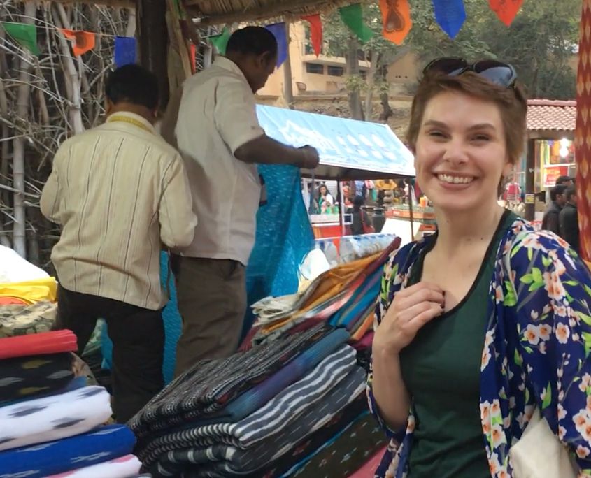 Young woman smiling at colorful fabric market stall with vendors in background.