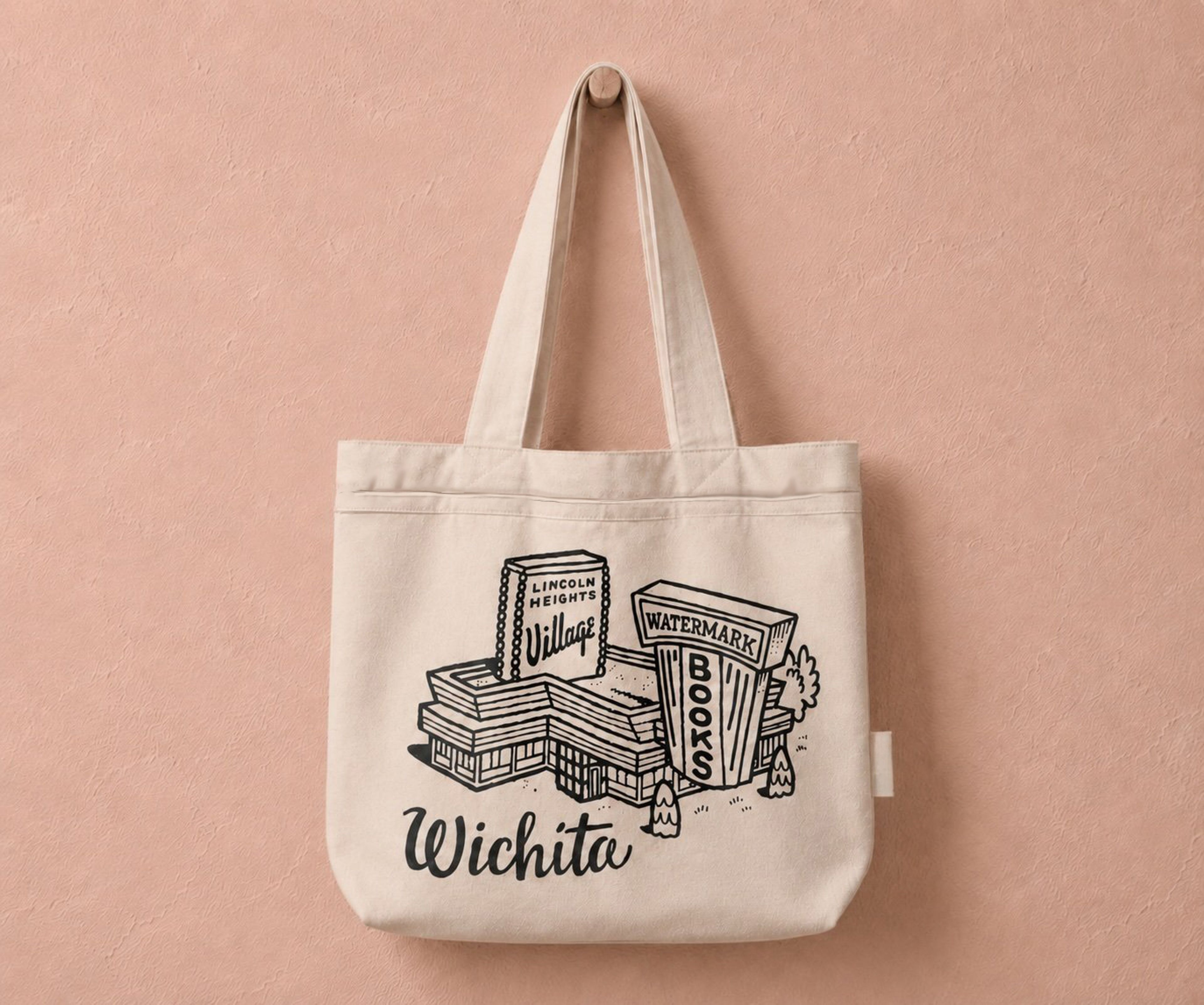 A canvas tote bag hanging on a weathered concrete wall, featuring black line-art illustrations of the Watermark Books & Cafe and Lincoln Heights Village buildings above the word "Wichita" in script.