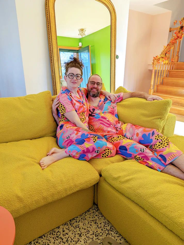 Two people in matching colorful pajamas sitting on a yellow couch in a bright home.