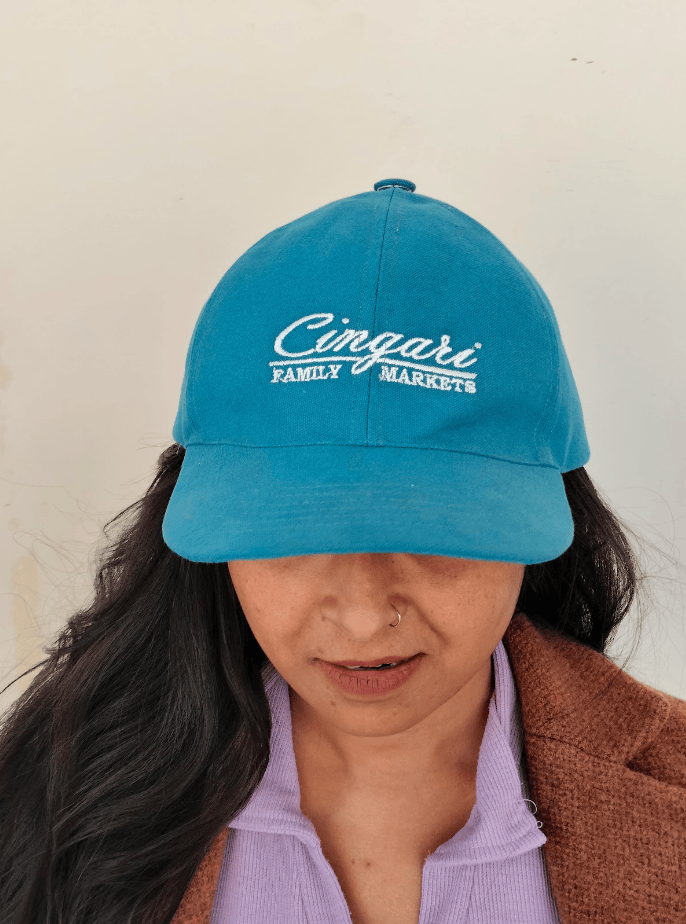 Woman wearing turquoise Cingari Family Markets baseball cap and brown cardigan.