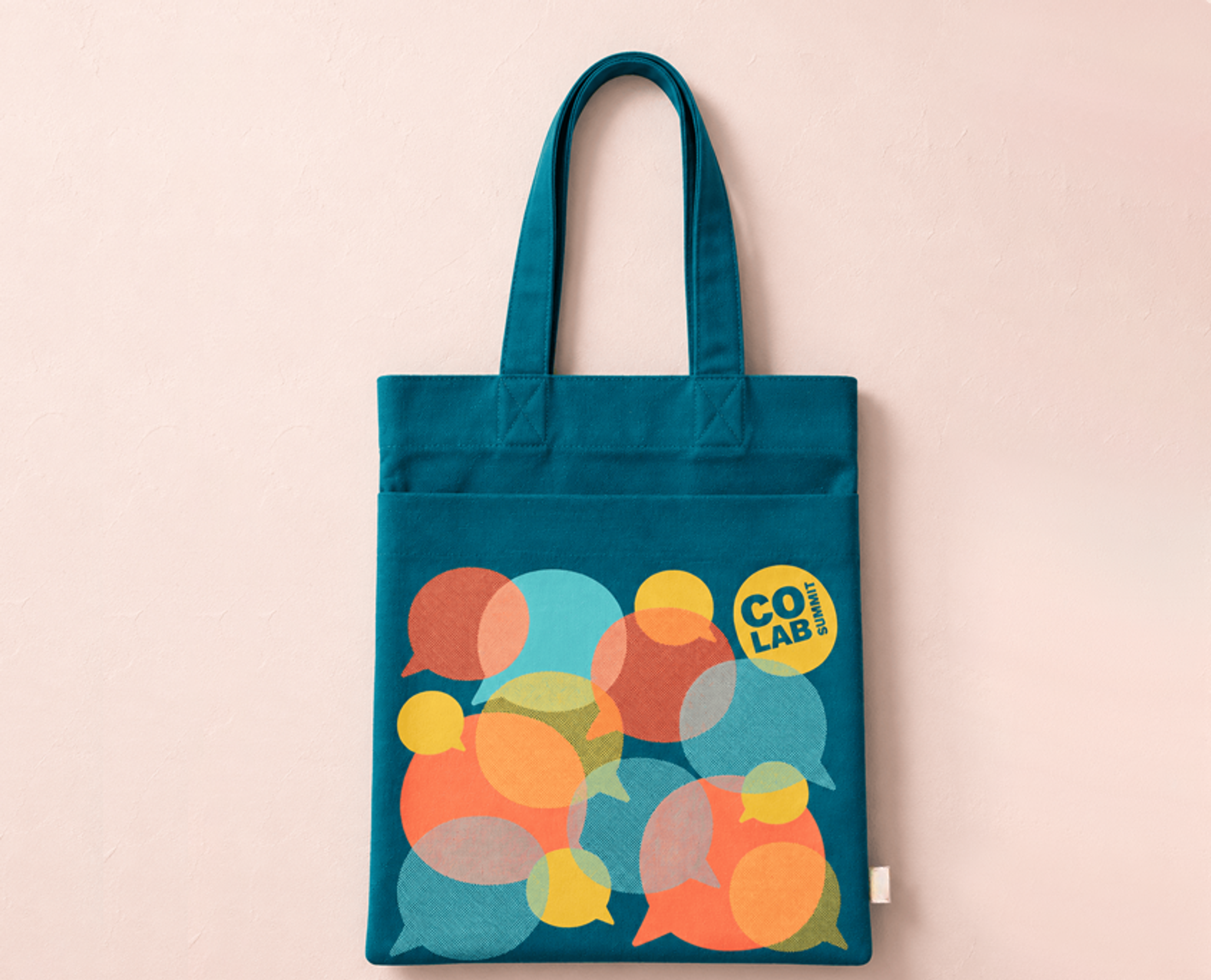 A teal canvas tote bag with colorful overlapping speech-bubble graphics hangs against a soft blush-pink plaster wall. The tote features long teal straps, a front pocket, and bold “CO LAB Summit” branding in yellow and navy.
