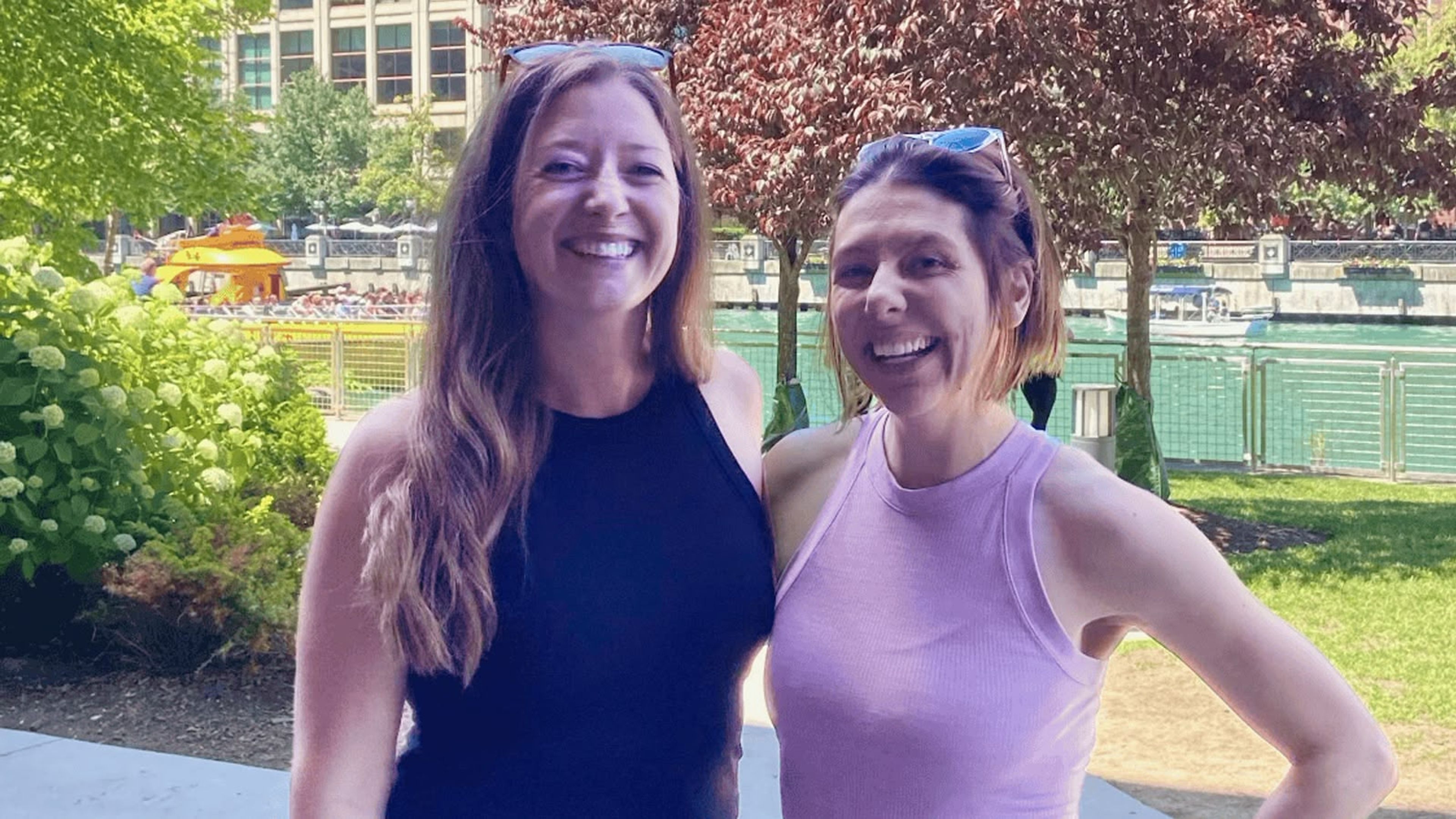 Two women smiling at camera in urban park with buildings and red foliage trees.