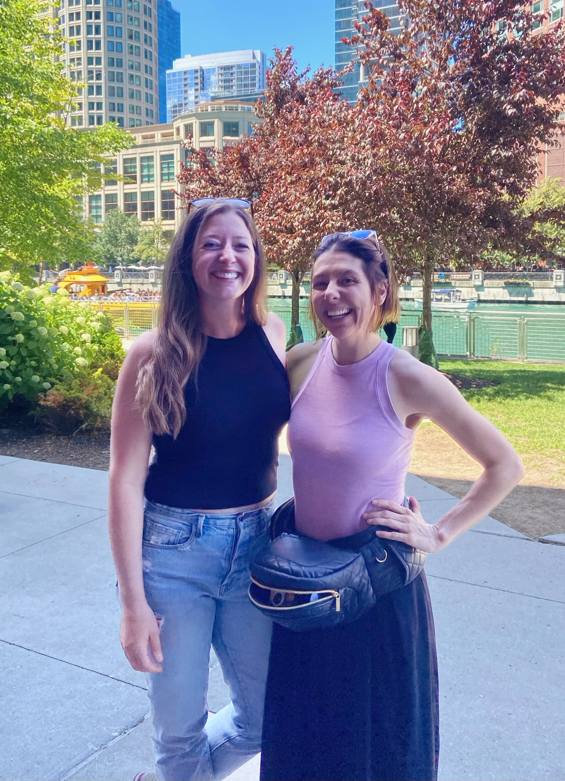 Two women smiling at camera in urban park with buildings and red foliage trees.