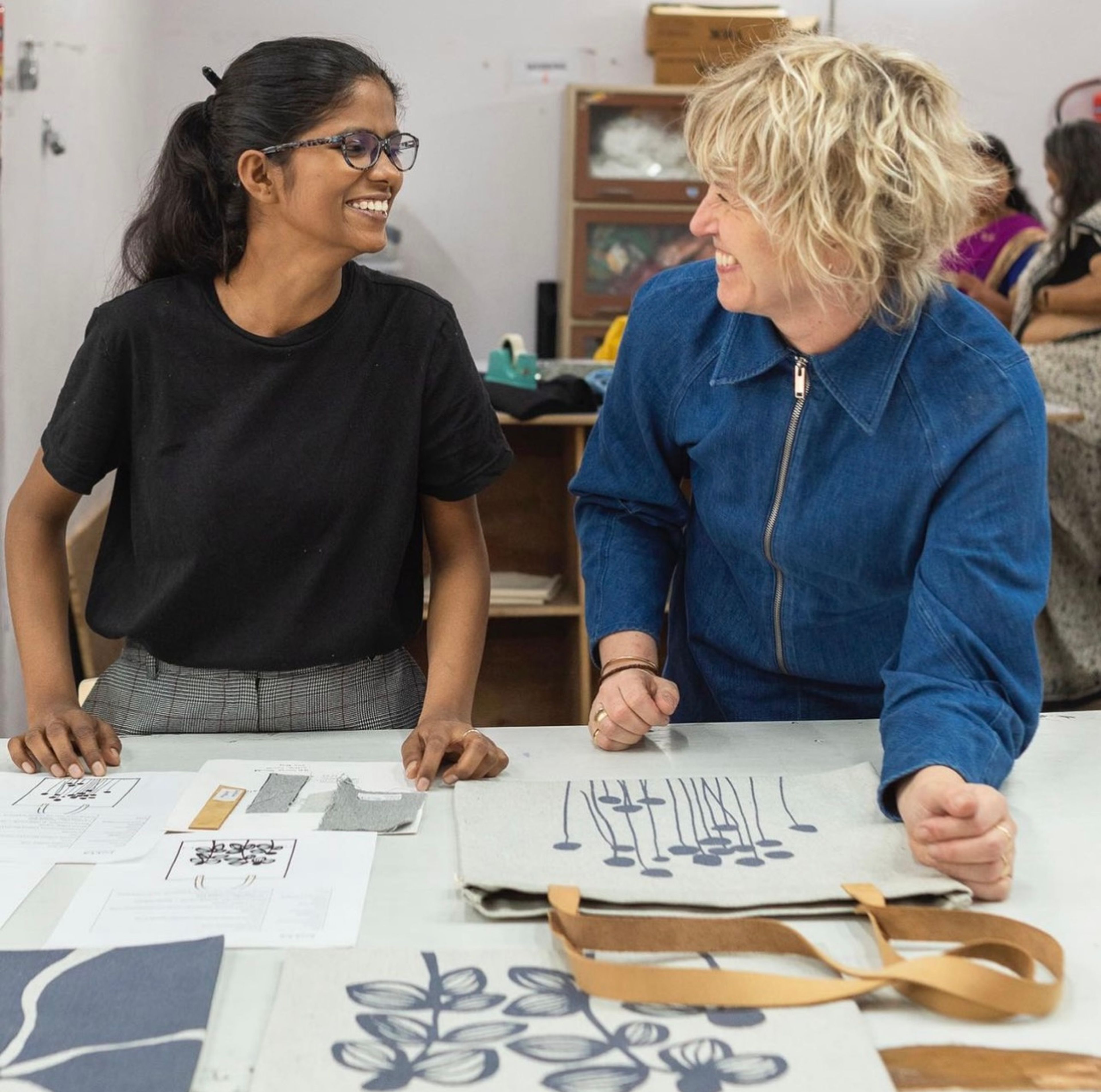Two women smiling while reviewing design sketches and fabric samples at a table.