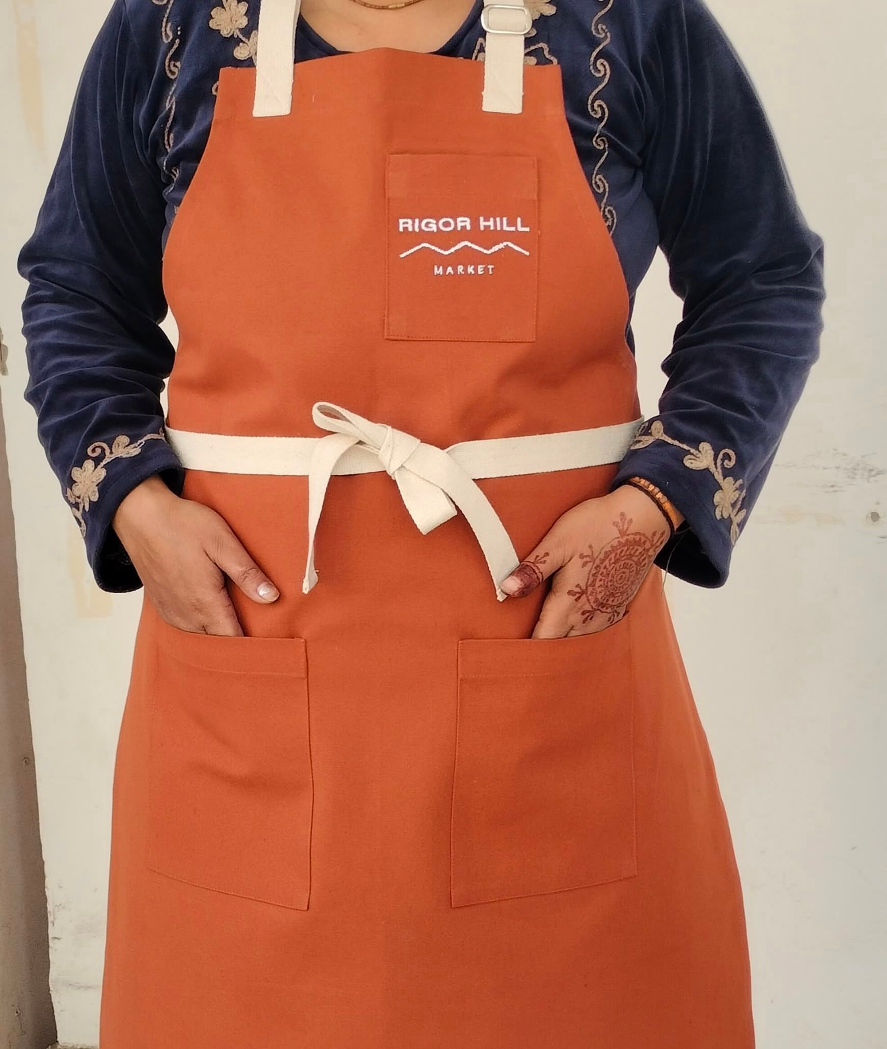 Person wearing orange Rigor Hill Market apron with white straps over navy embroidered jacket.