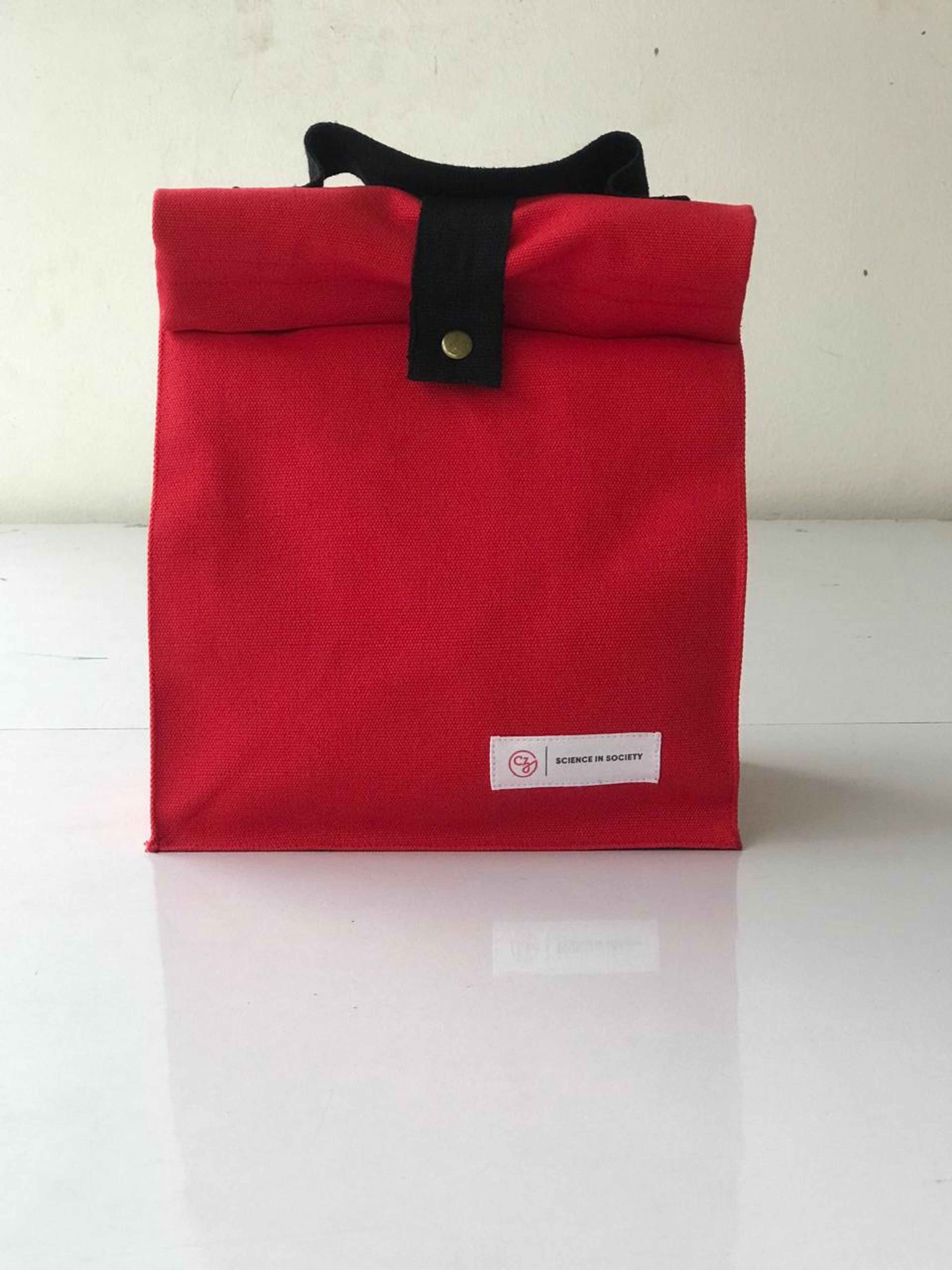 Red fabric lunch bag with black handle and "Science in Society" label.