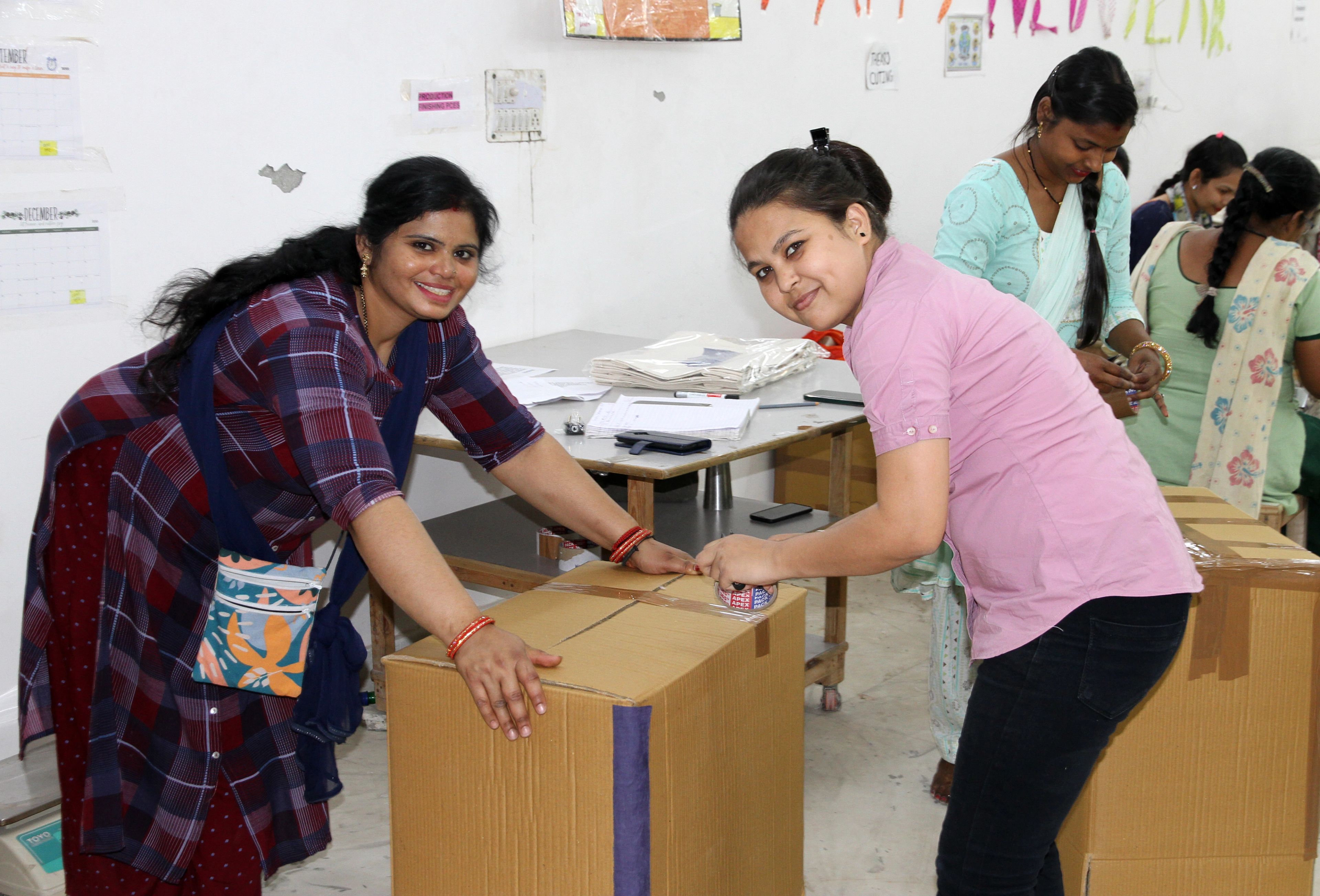Woman in pink shirt sealing cardboard box while colleague in plaid shirt watches in classroom.
