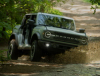 A dark green Ford Bronco splashes through a mud puddle on a dirt road in the forest.