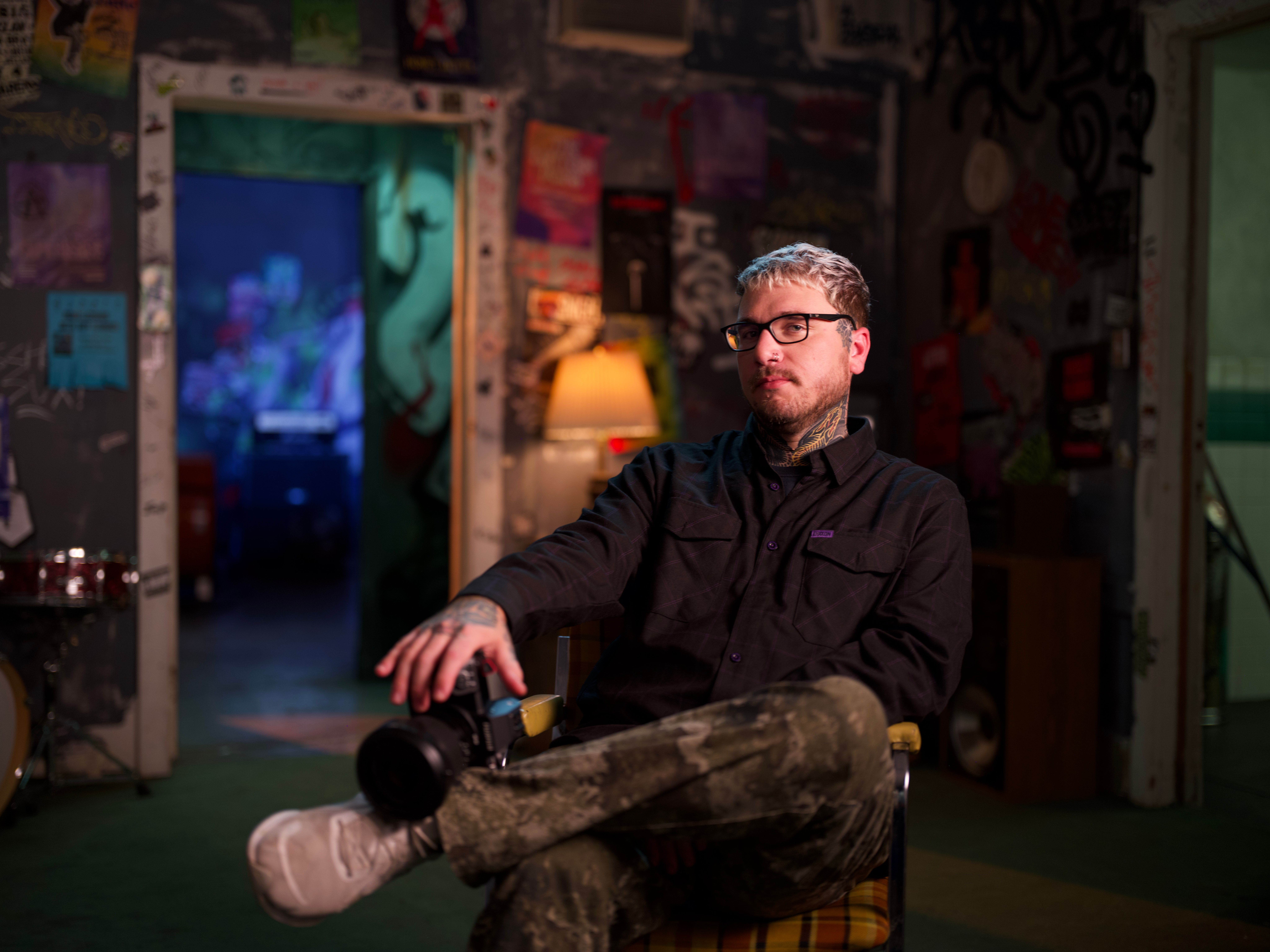 A tattooed man with light hair and glasses sits holding a camera in an eclectic room covered with posters and graffiti, with a blue-lit doorway in the background.