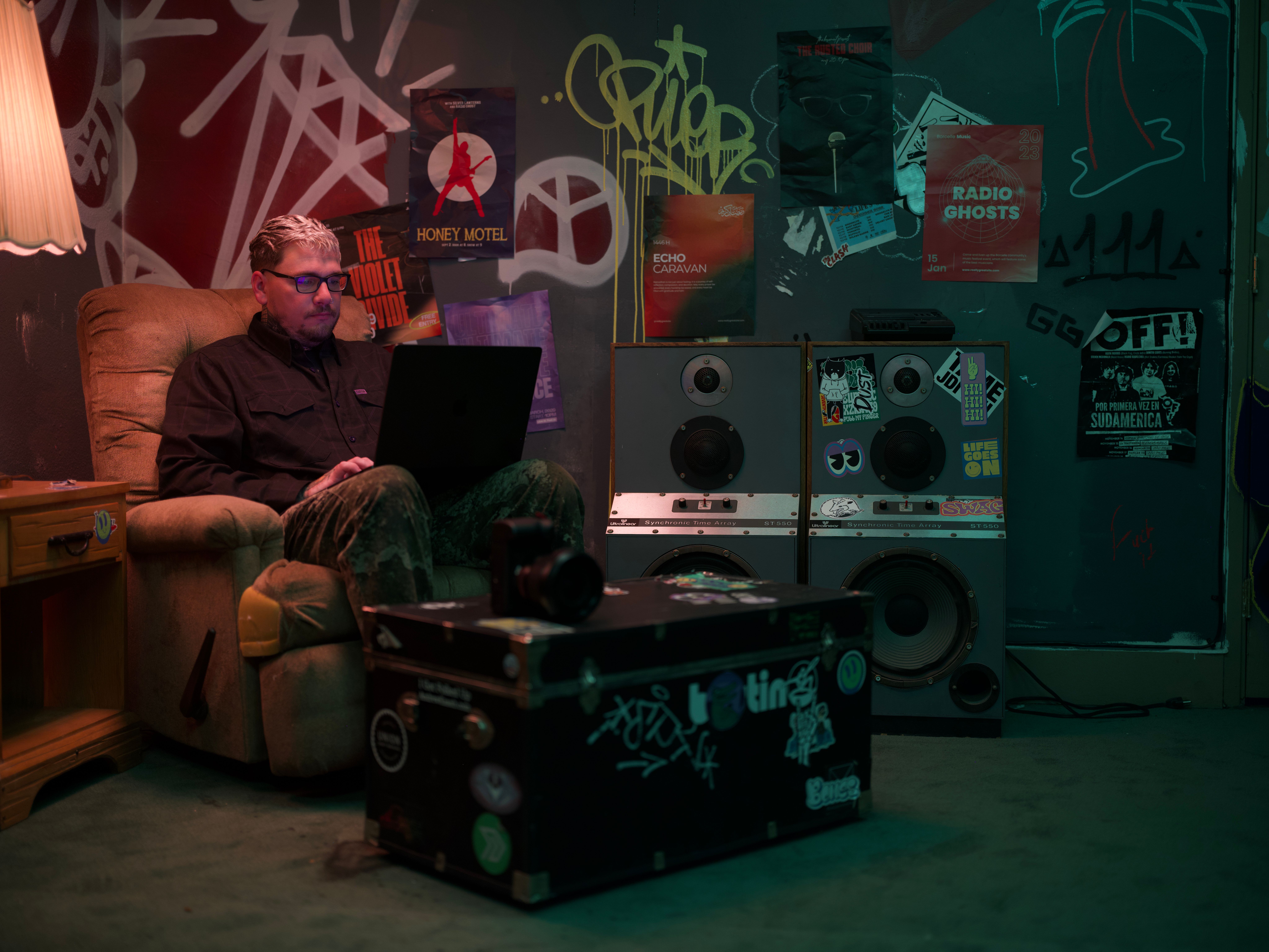 A man with glasses sits in a recliner, typing on a laptop, in a dimly lit room with graffiti-covered walls, posters, and large speakers.