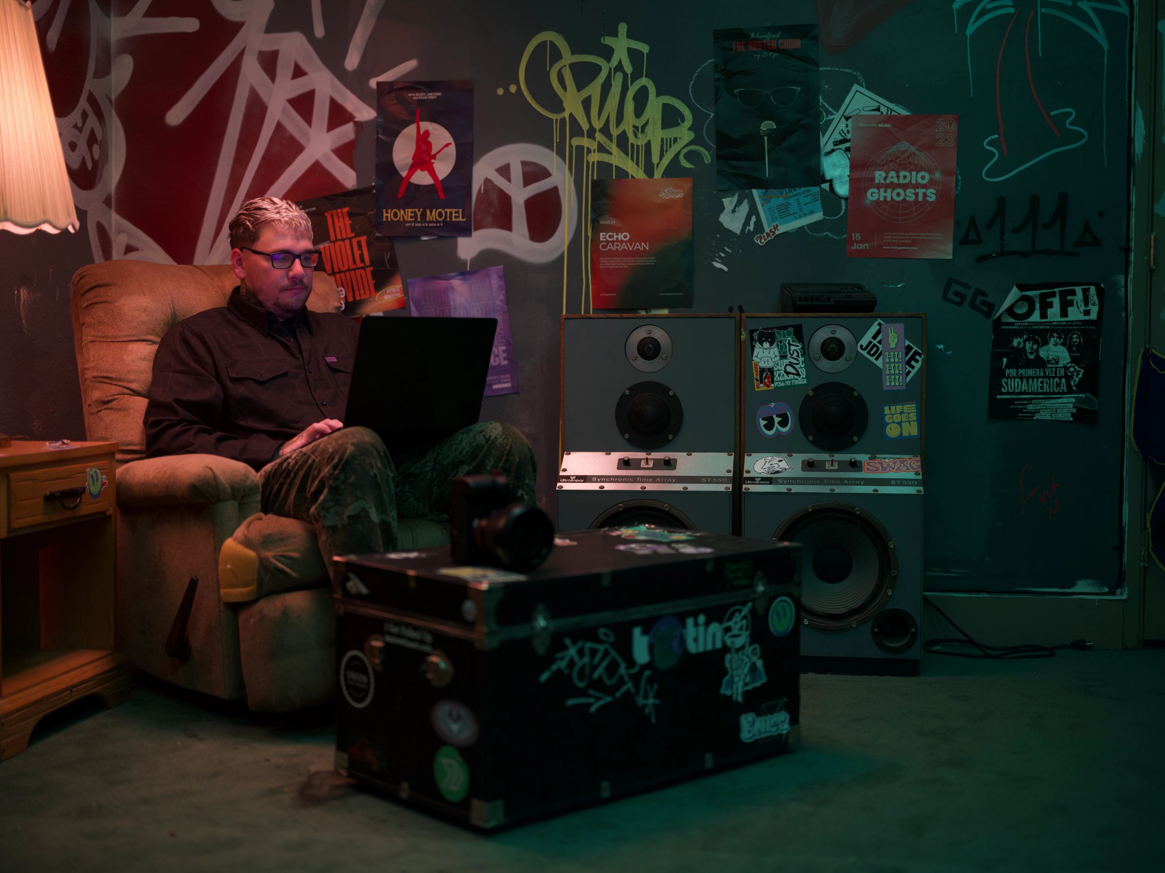 A man with glasses sits in a recliner, typing on a laptop, in a dimly lit room with graffiti-covered walls, posters, and large speakers.