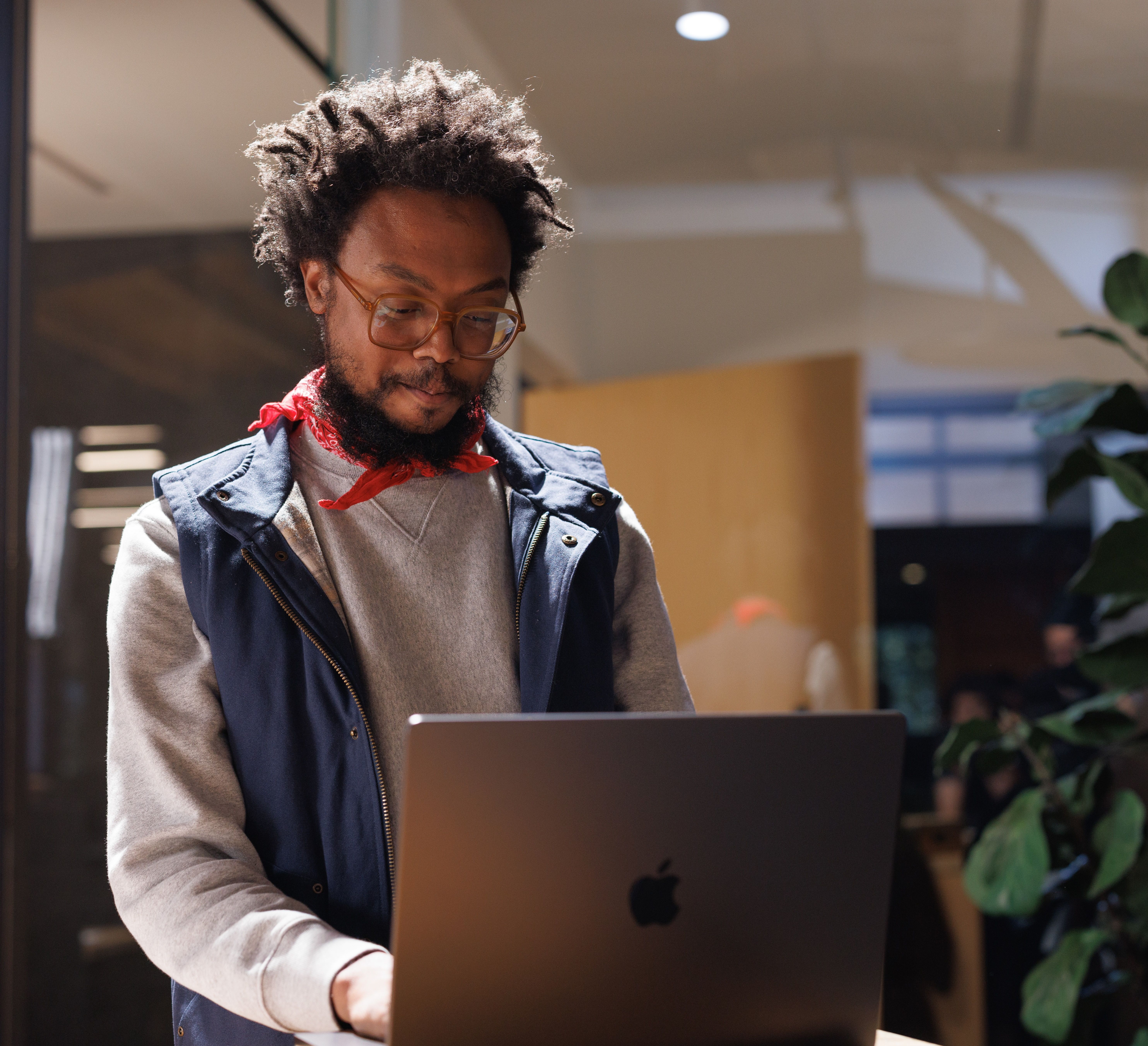 A Black man with dreadlocks, glasses, and a beard, wearing a red bandana and blue vest, looks at an open Apple laptop.