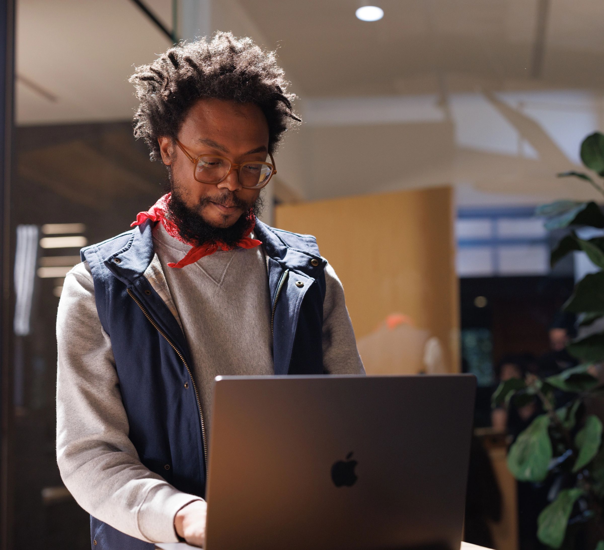 A Black man with dreadlocks, glasses, and a beard, wearing a red bandana and blue vest, looks at an open Apple laptop.