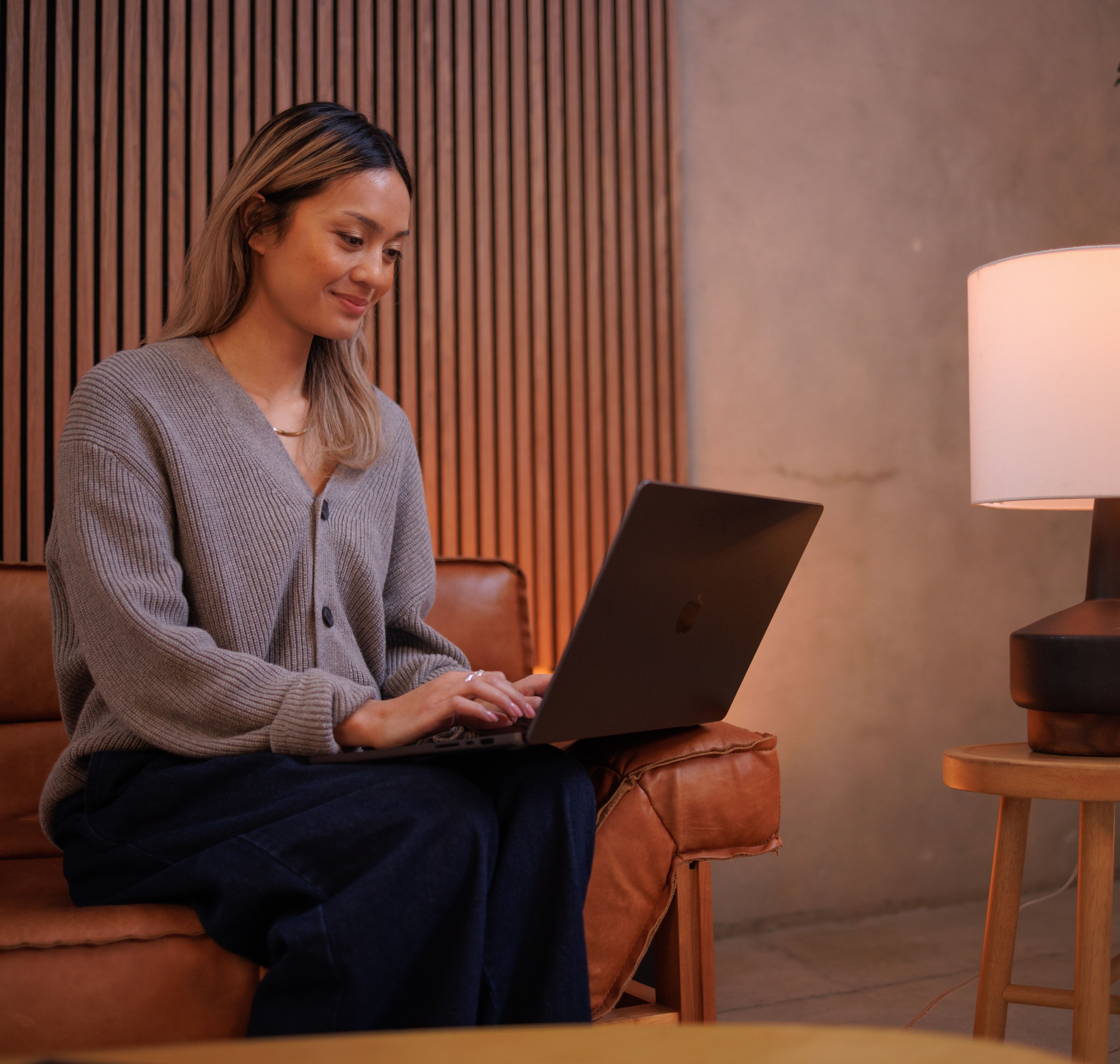 A smiling woman sits on a couch, working on a laptop.