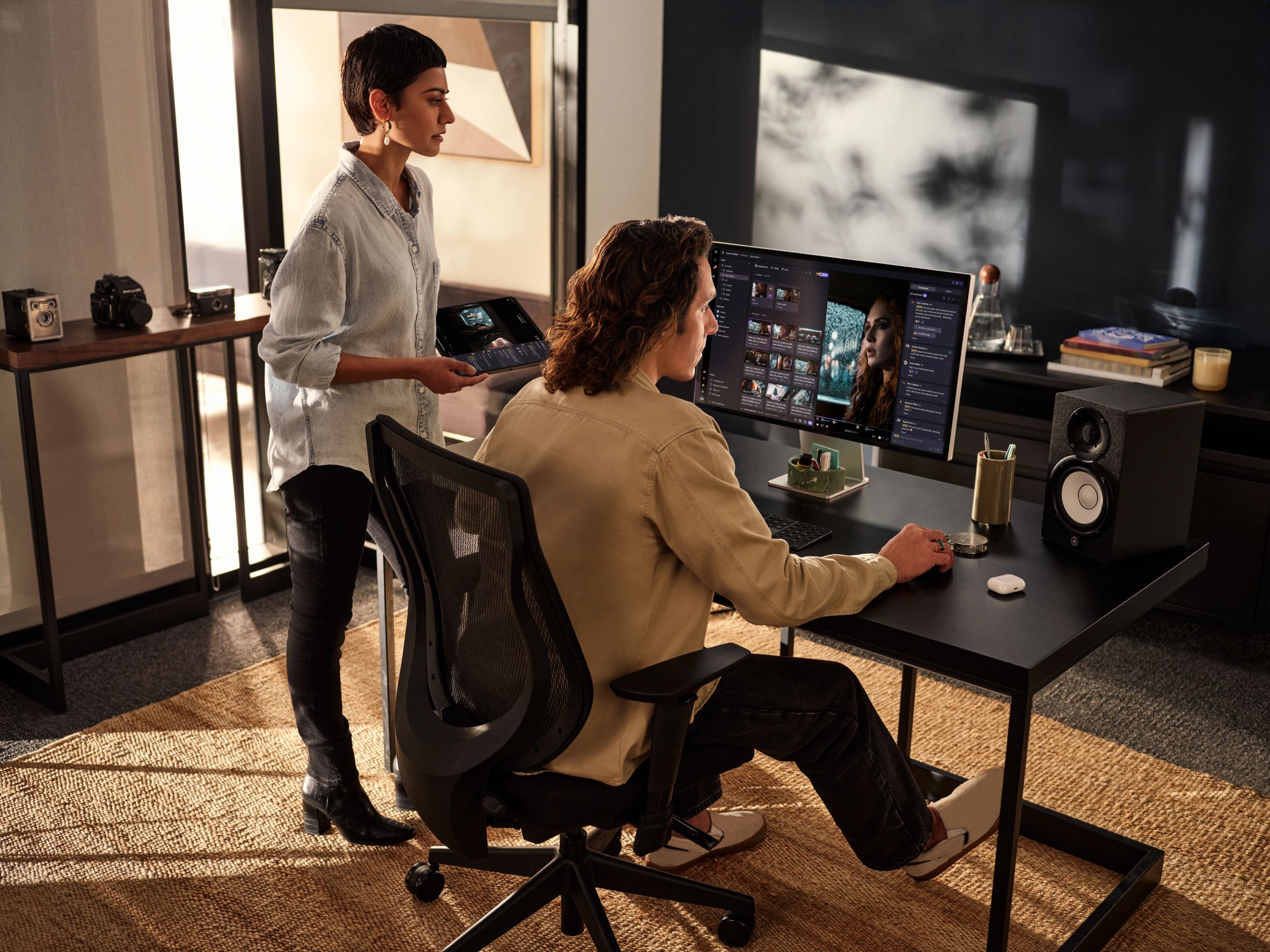 A man edits photos on a large monitor at a desk, while a woman stands behind him holding a tablet.