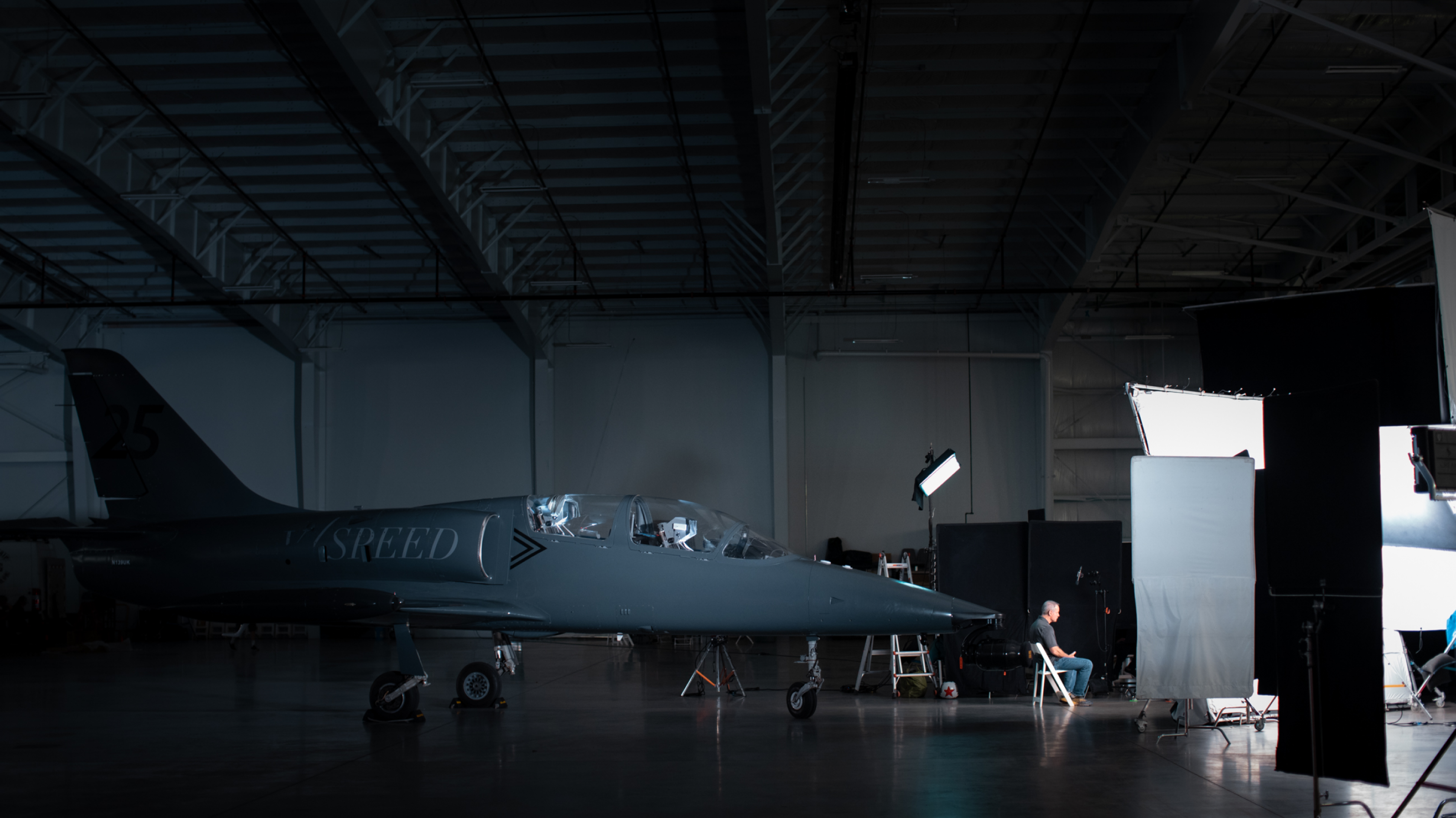 Man sitting in a hangar with a dark gray jet behind him. He is being interviewed.